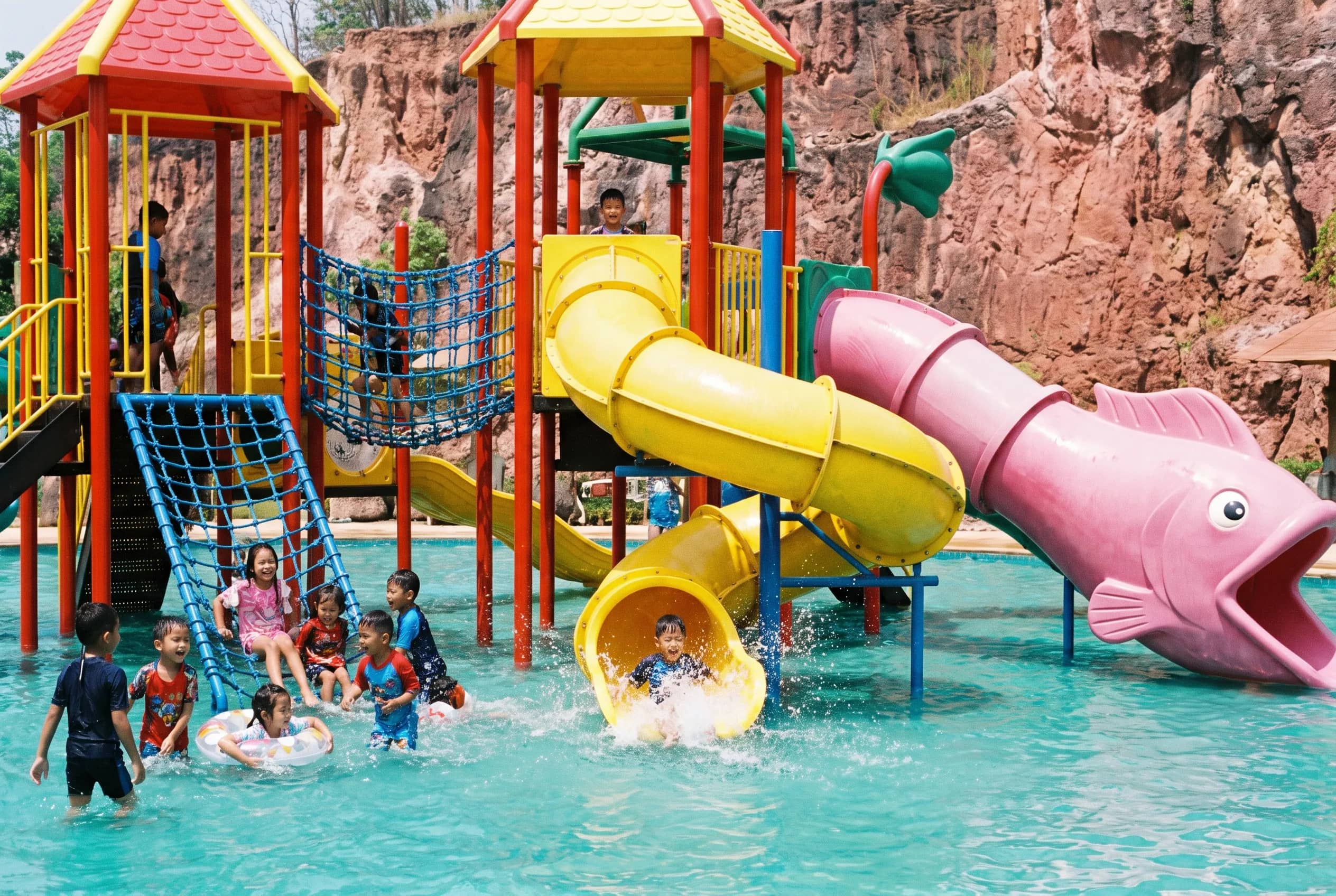 Kids play in a turquoise pool beside a big pink fish slide and red and yellow towers