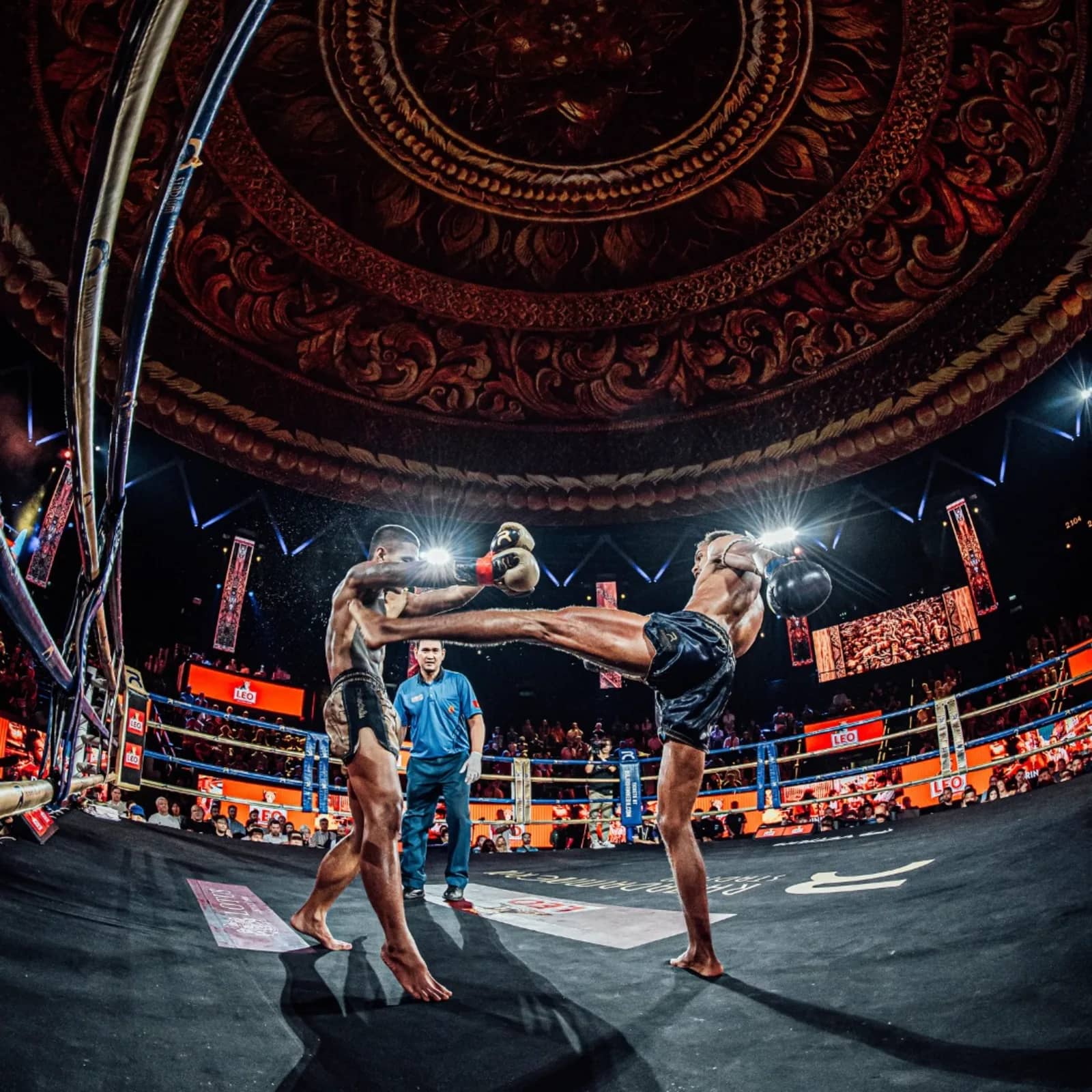 Two fighters exchange kicks in the ring as a referee watches beneath a decorated dome ceiling