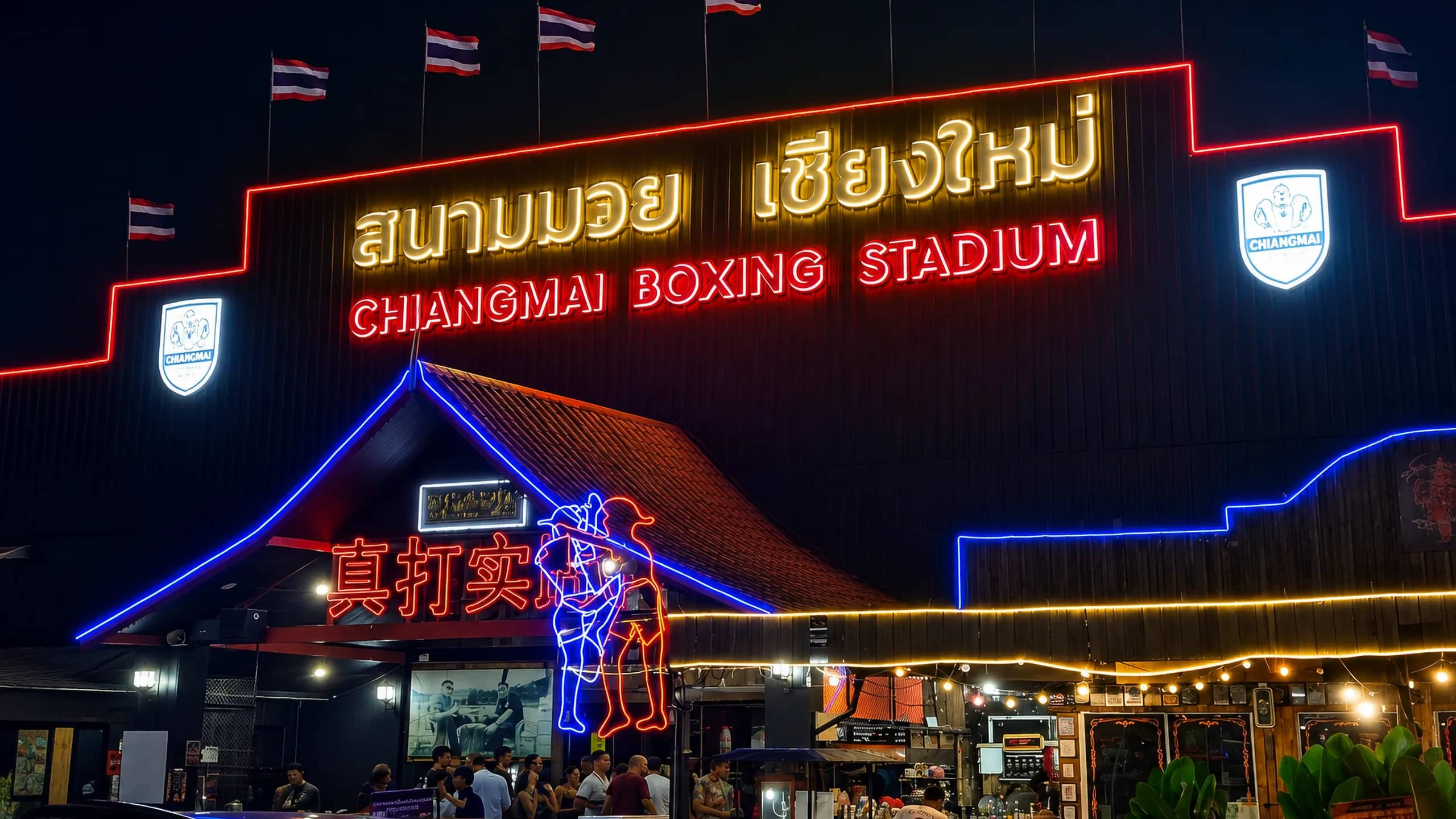 The neon-lit entrance sign of Chiang Mai Boxing Stadium at night with flags along the roof