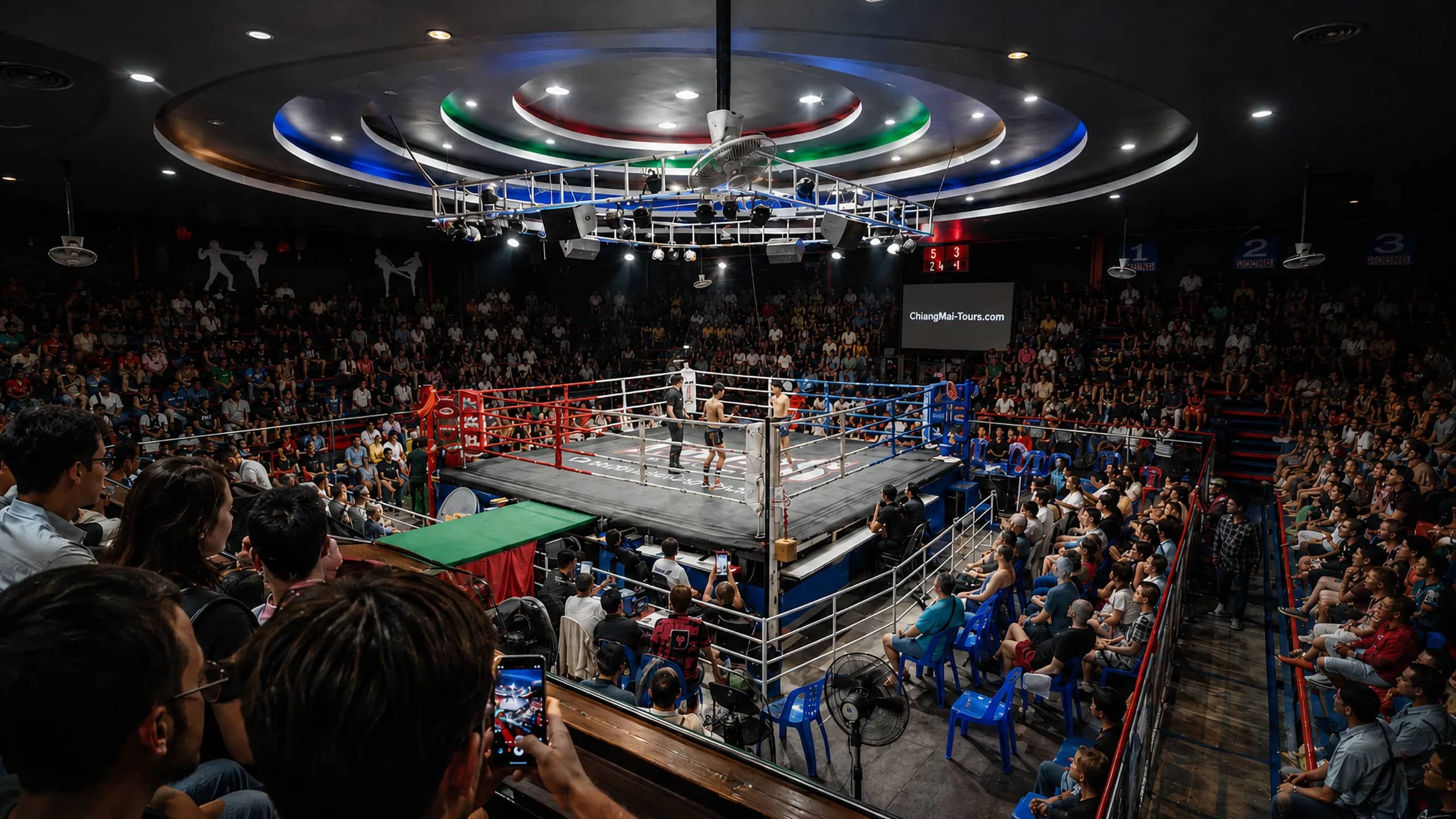 Spectators fill the seats around the ring under circular ceiling lights at Chiang Mai Boxing Stadium