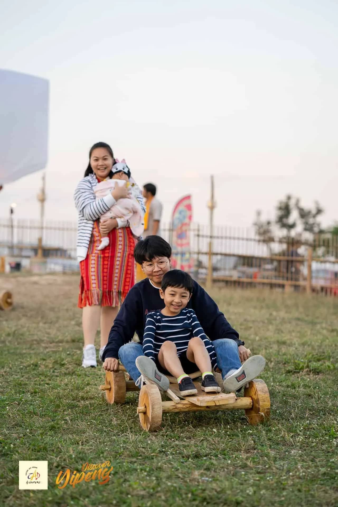 Children ride a wooden go-kart across the grass as their mother watches at the festival grounds