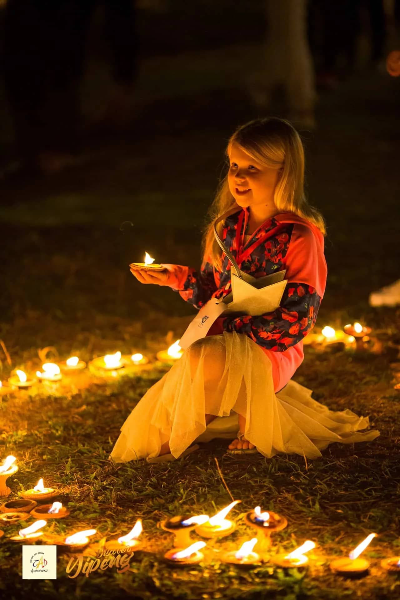 A girl in a festive dress holds a small candle among rows of glowing oil lamps on the grass