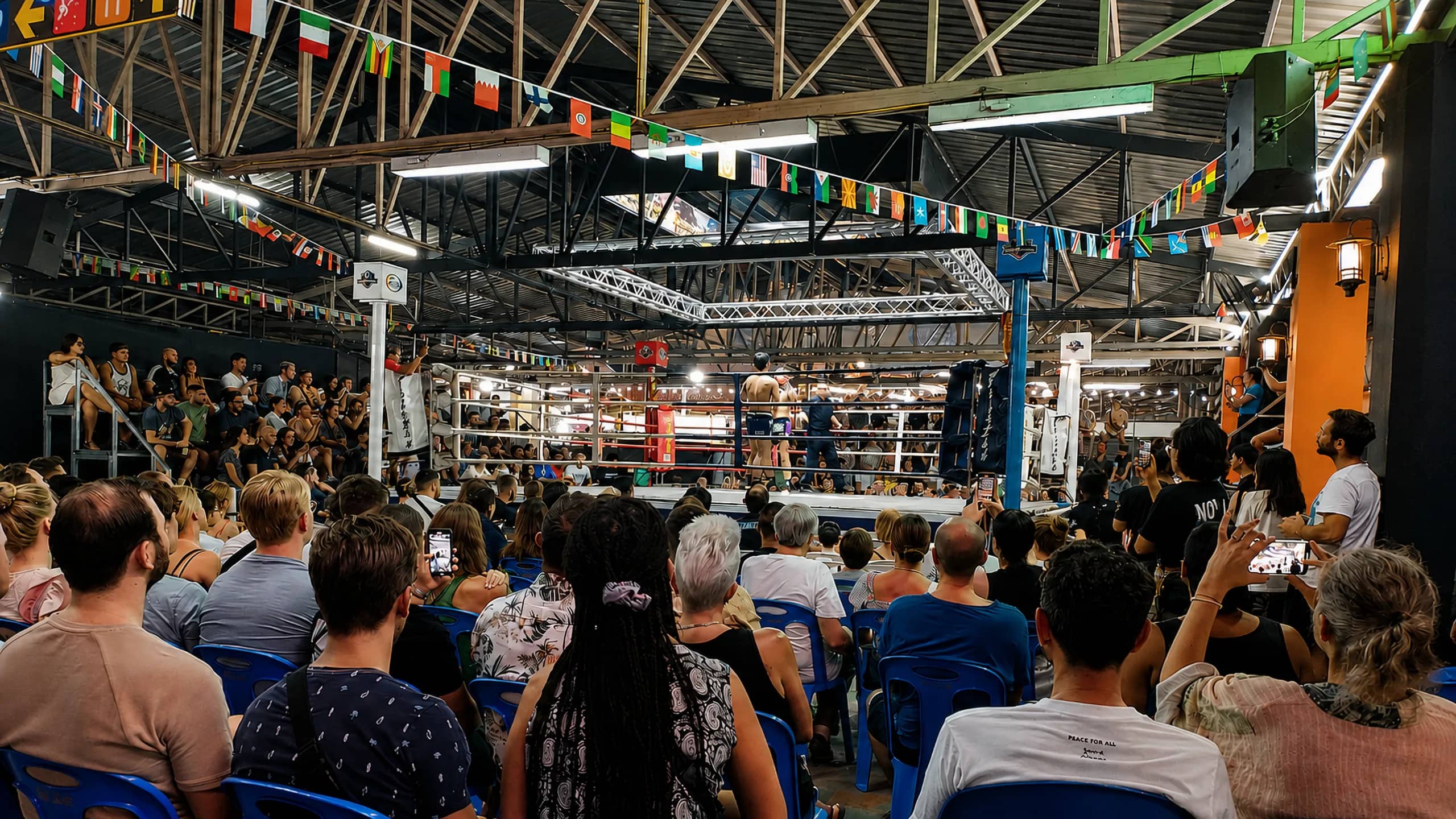 A large crowd under strings of flags watches a Muay Thai bout at the Night Bazaar boxing stadium