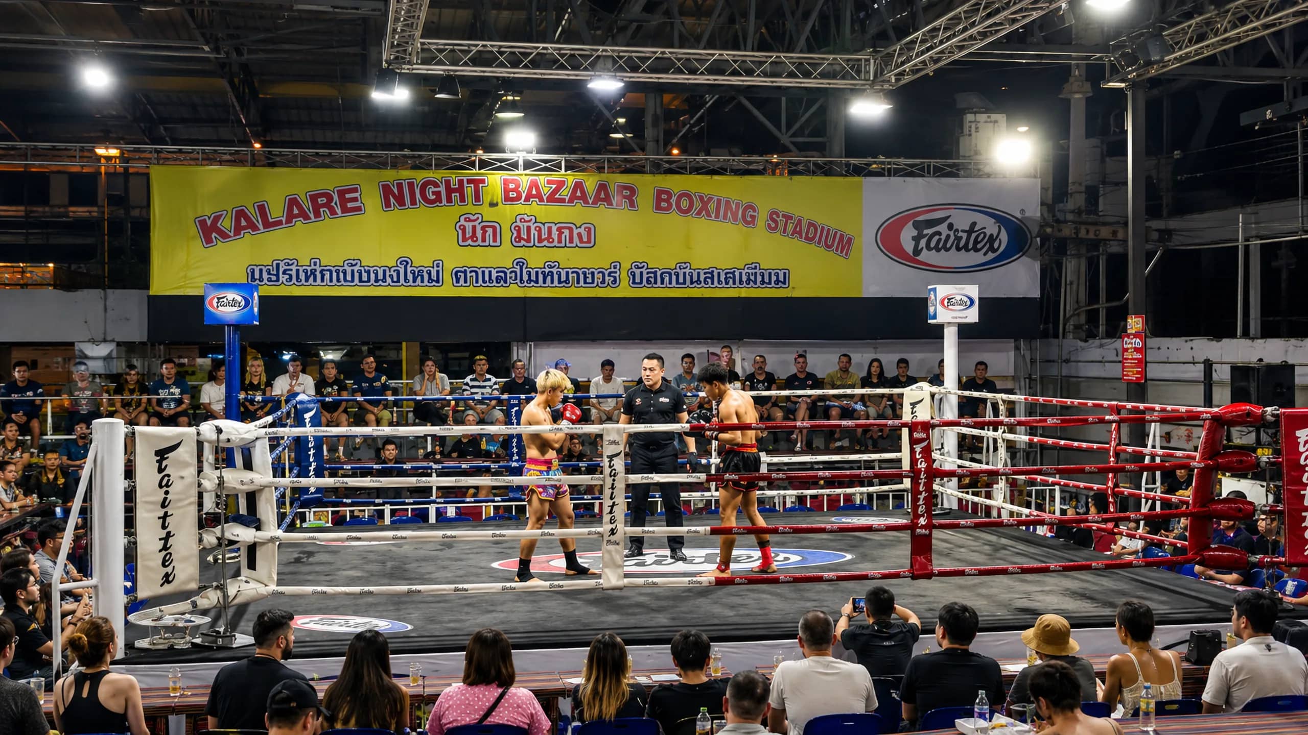 Fans sit around the ring as two fighters face off under bright lights at the Night Bazaar boxing stadium