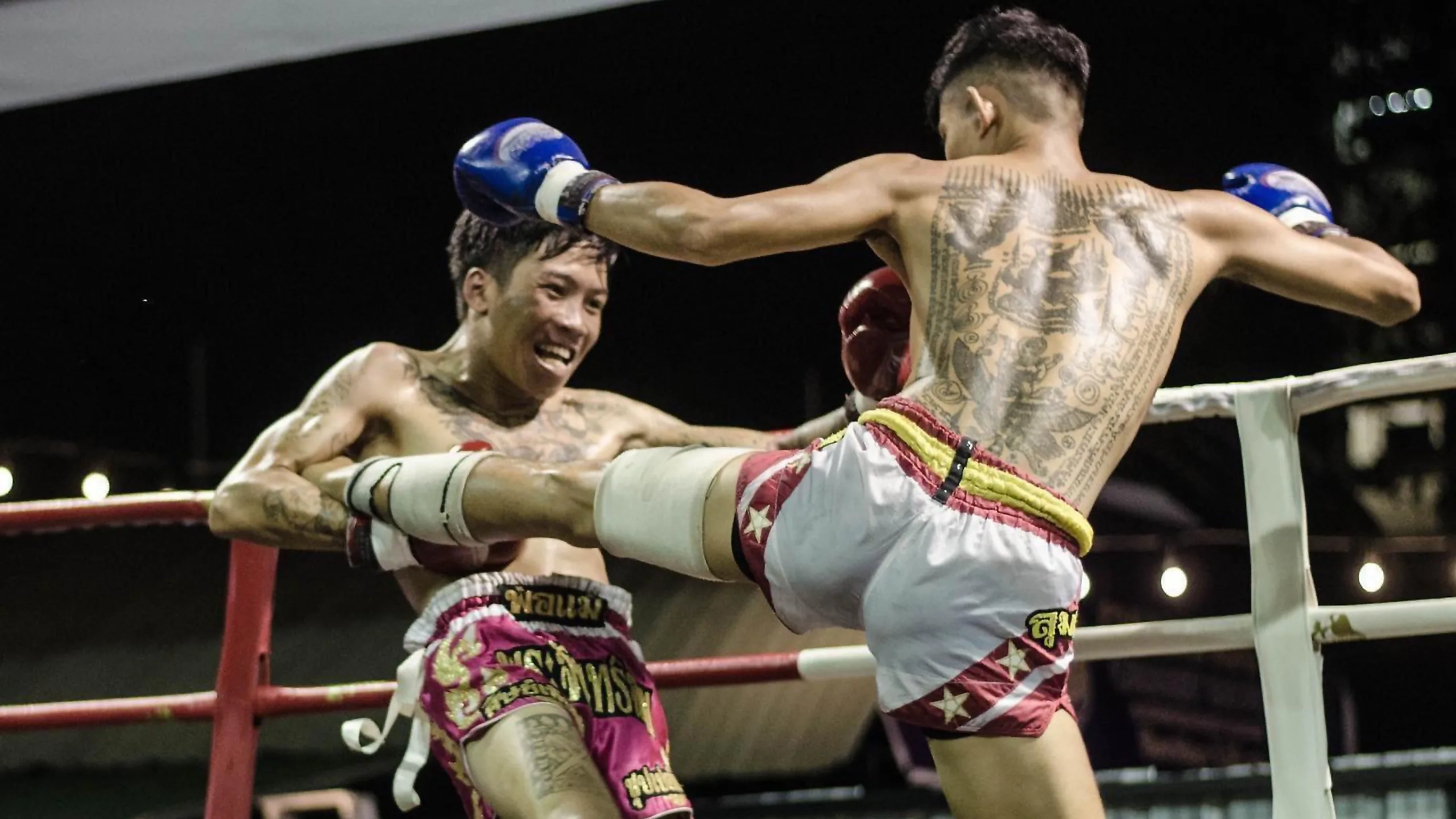 A tattooed fighter throws a high kick at his opponent against the ropes in Chiang Mai