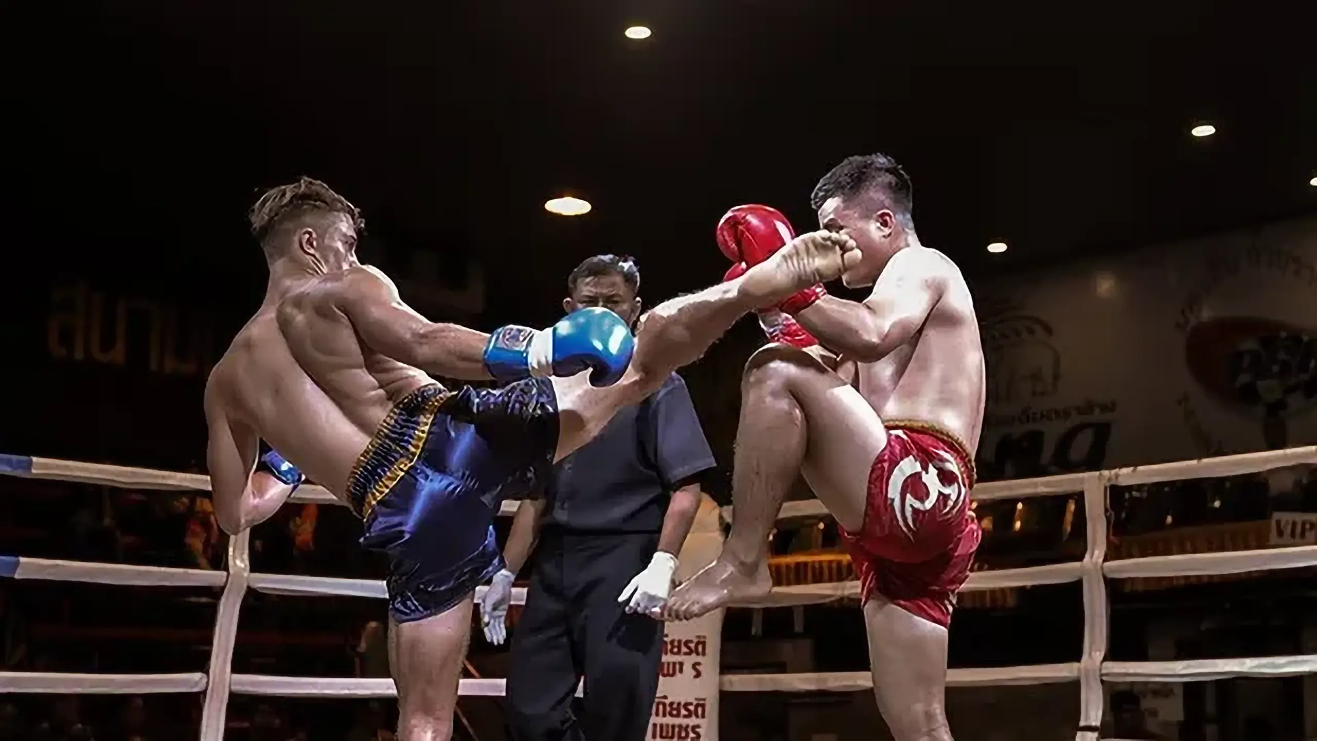 Two Muay Thai fighters throw kicks at each other in the ring while the referee looks on