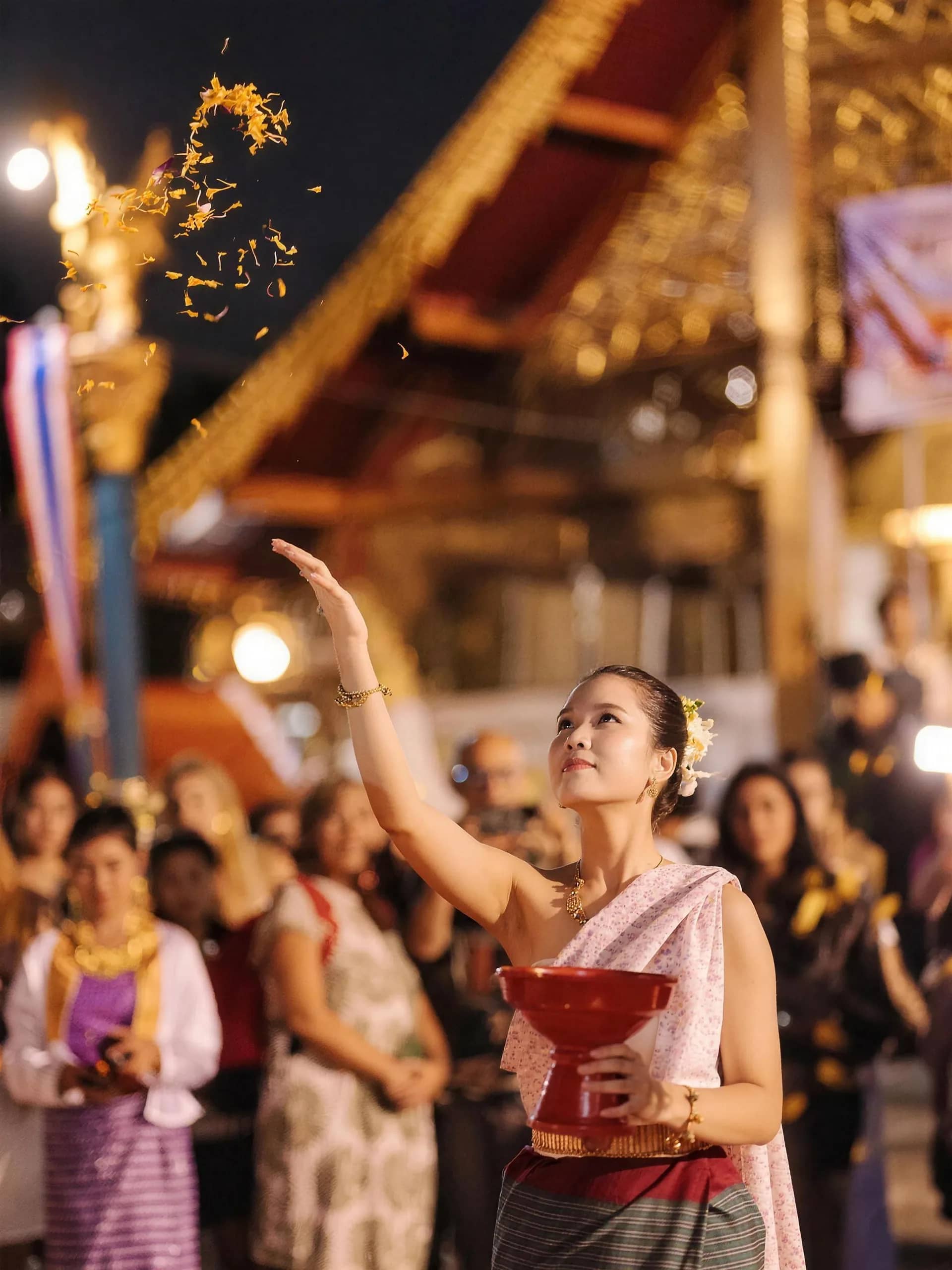 A woman in traditional Lanna dress tosses petals from a bowl in front of a festival crowd at night