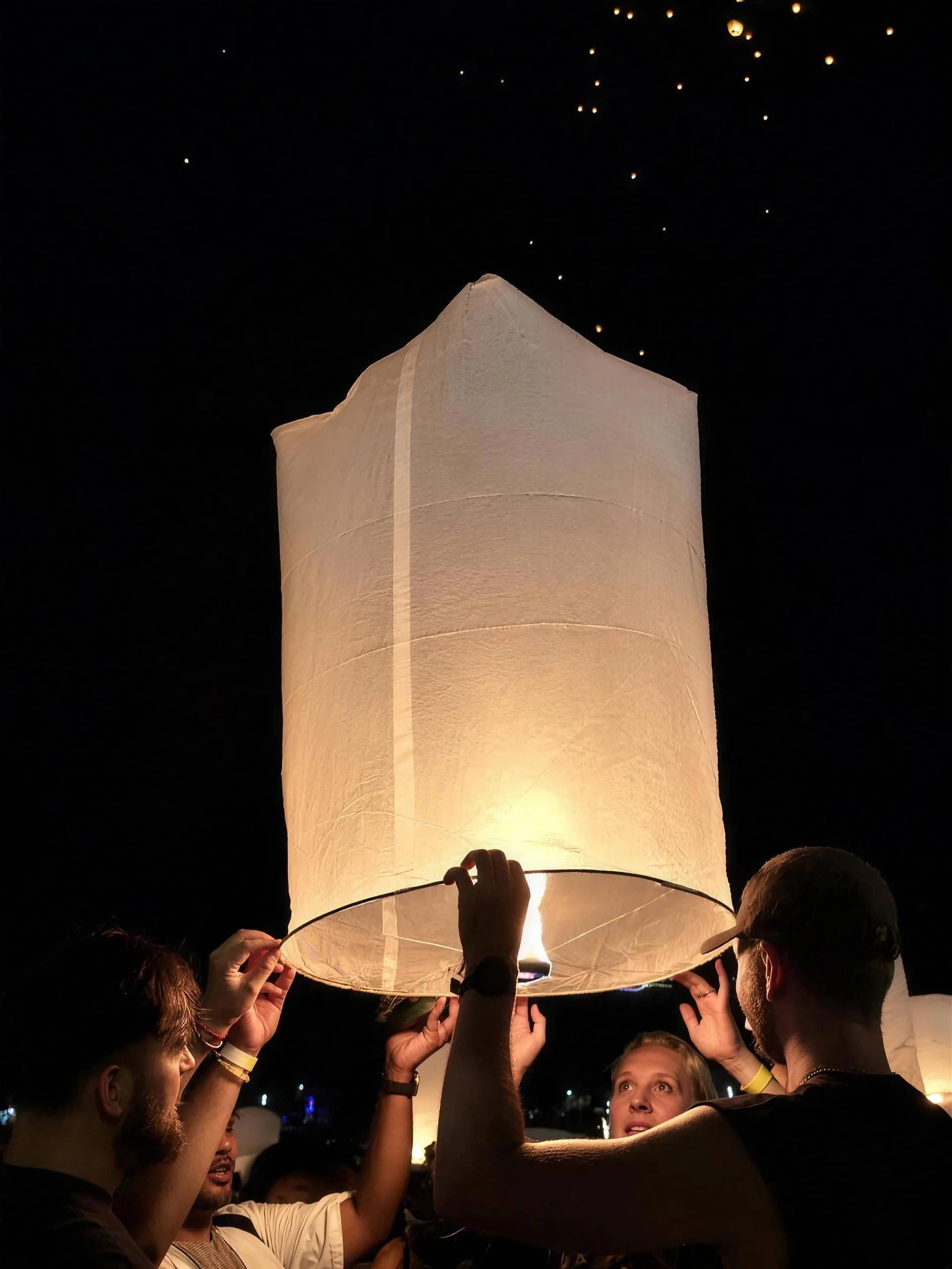 Hands hold up a large white sky lantern lit from below as other lanterns drift overhead