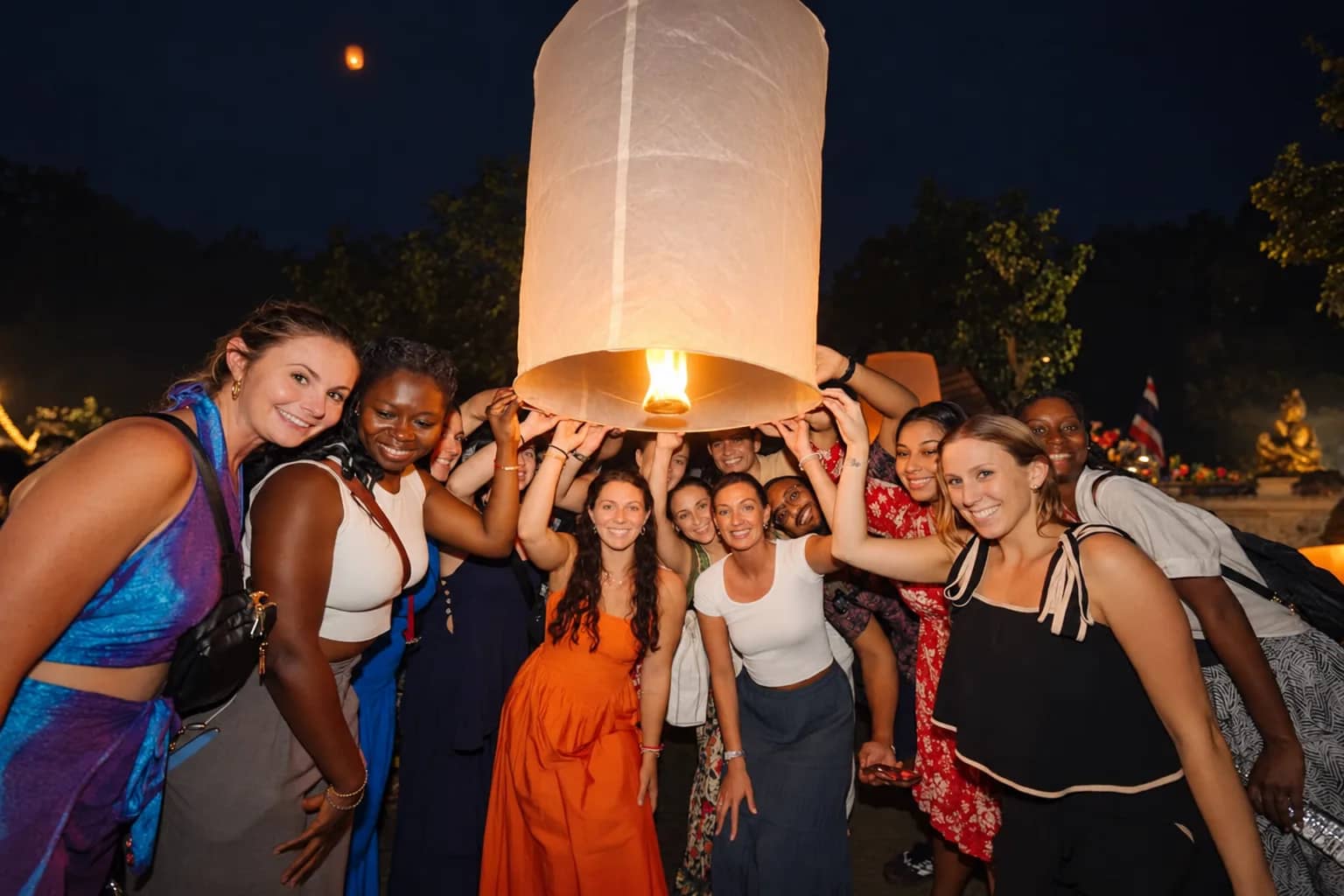 A smiling group of guests gathers under a large glowing paper lantern at the Doi Saket festival
