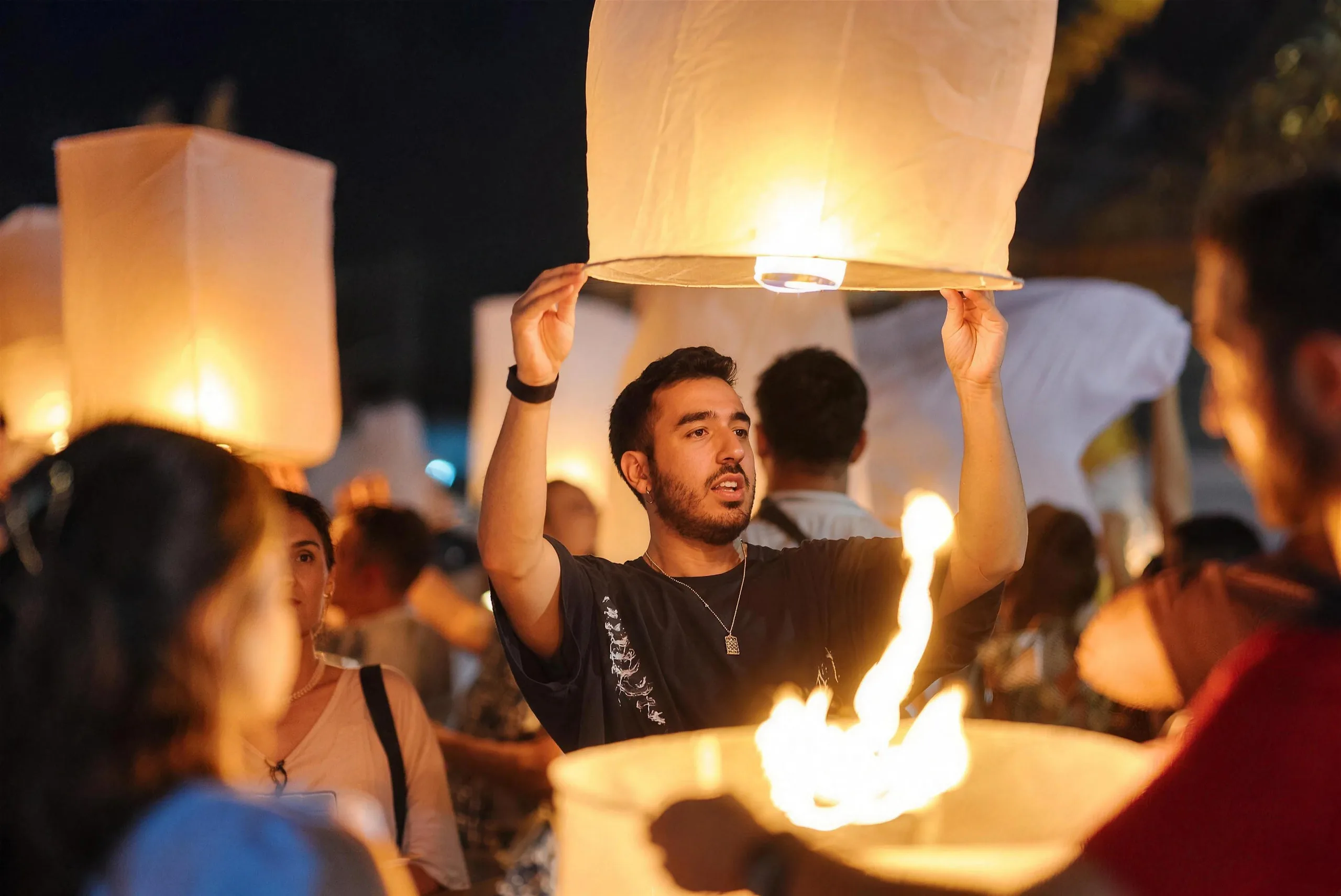 A man raises a glowing paper sky lantern above a crowd of festival guests at night