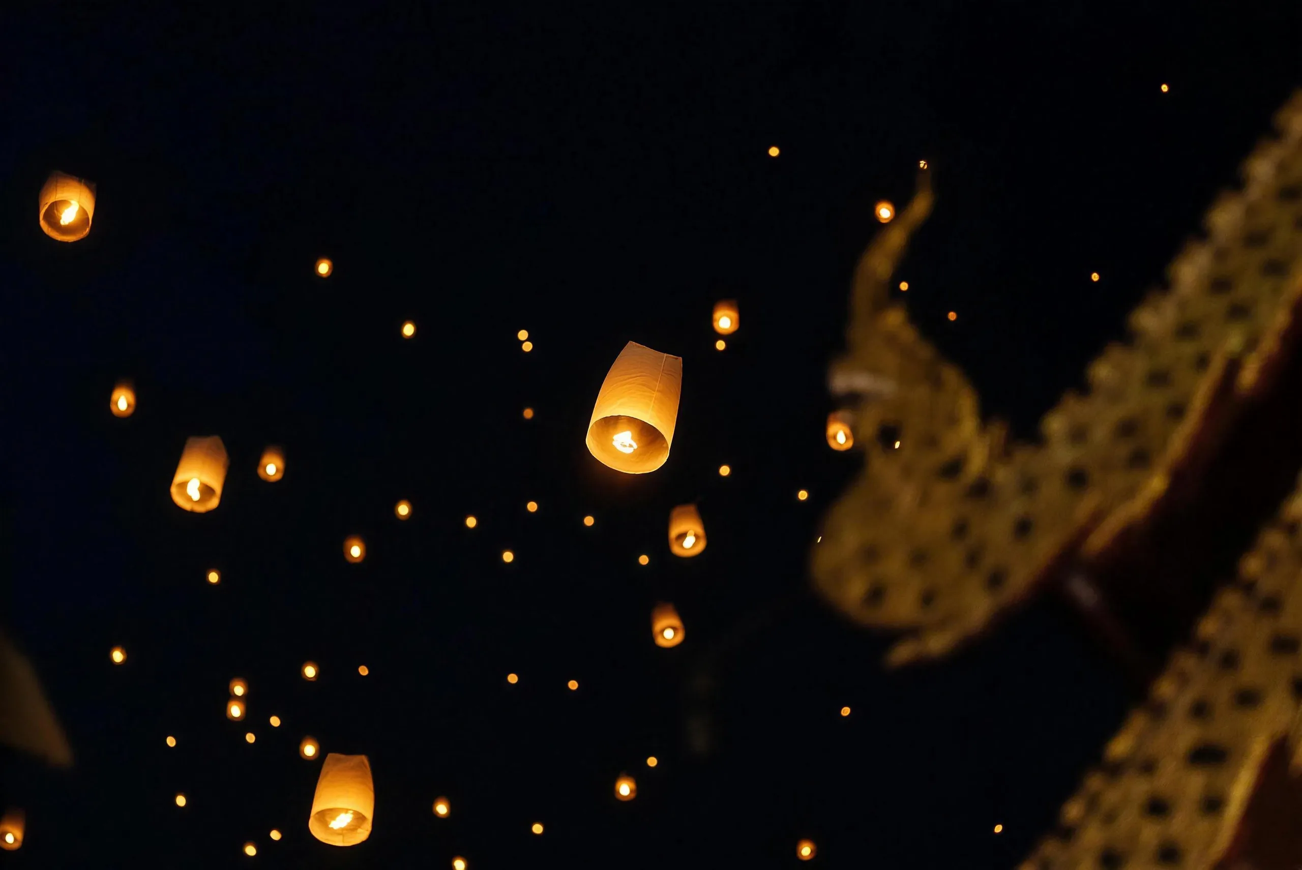 Glowing sky lanterns float in the night sky beside the golden gable of a temple roof