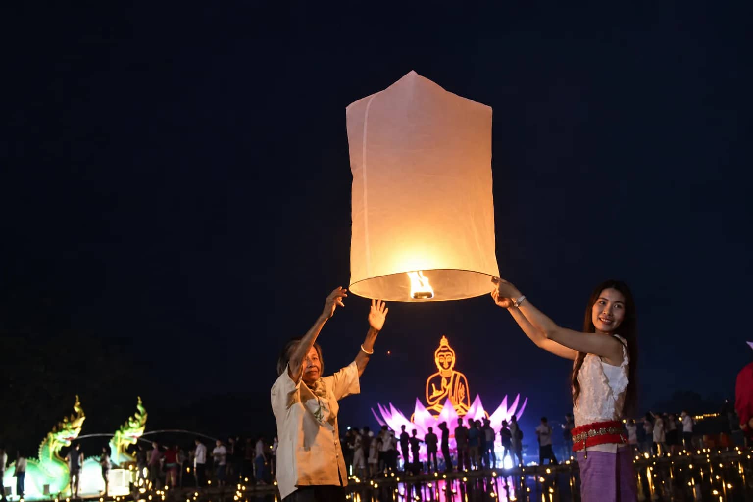 Two people lift a lit sky lantern with an illuminated golden Buddha and crowd behind them