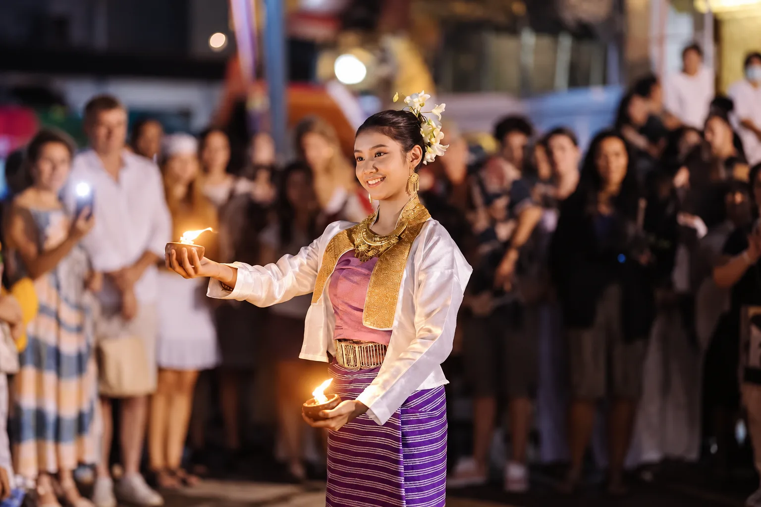 A woman in Lanna costume holds small clay oil lamps as festival guests watch behind her