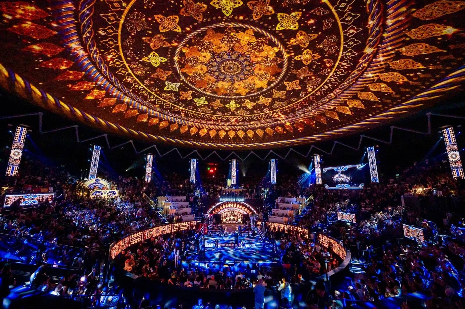 A packed Muay Thai stadium under an ornate carved dome with a lit ring in the centre