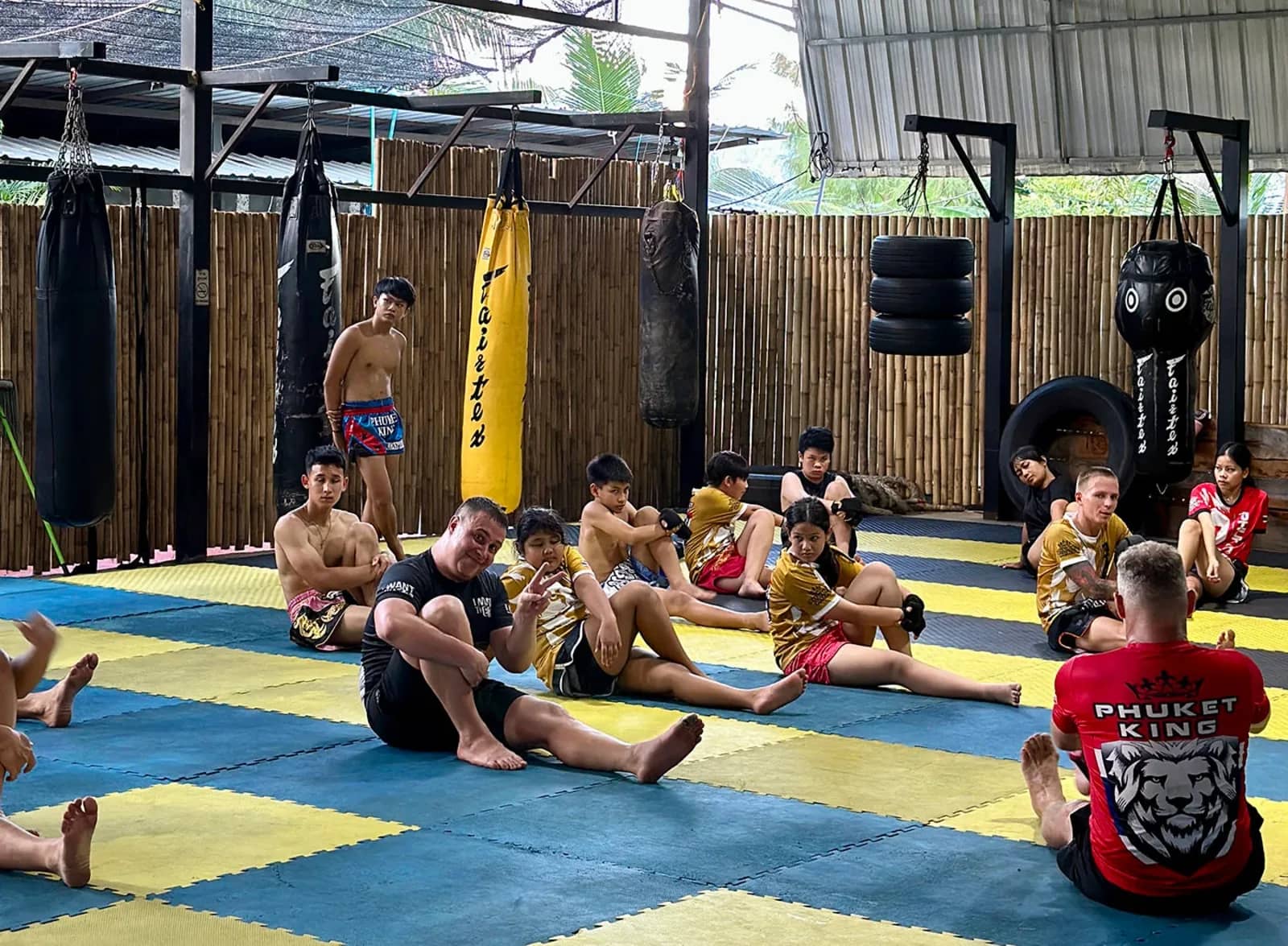 Students sit on blue and yellow mats stretching after a training session among hanging punching bags