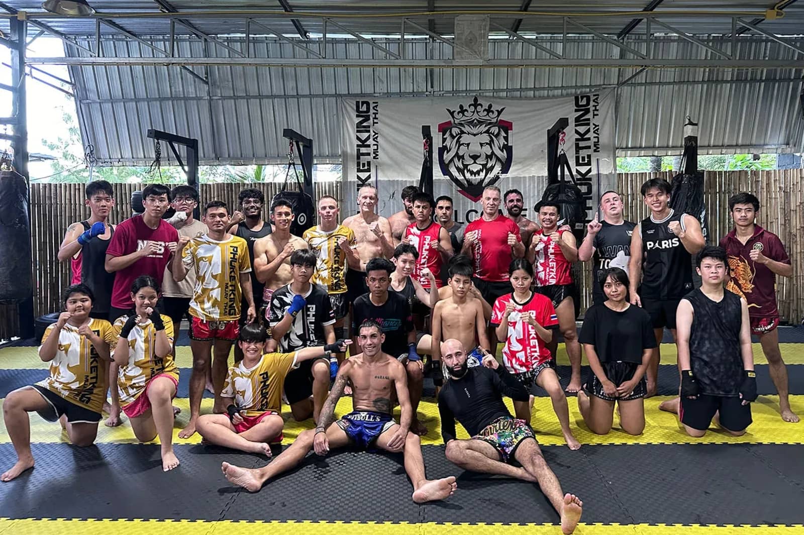 A large group of trainees and trainers poses on the mats under a Phuket King Muay Thai banner