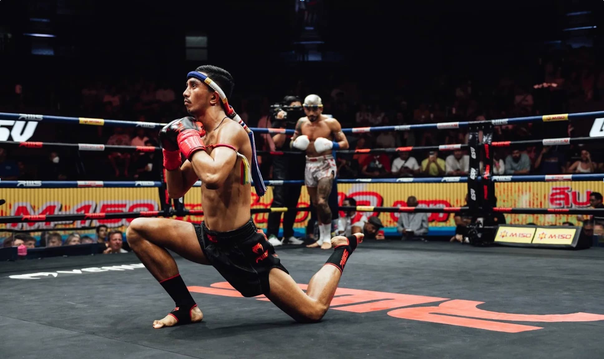A Muay Thai fighter in a headband kneels in the ring performing the pre-fight wai kru dance