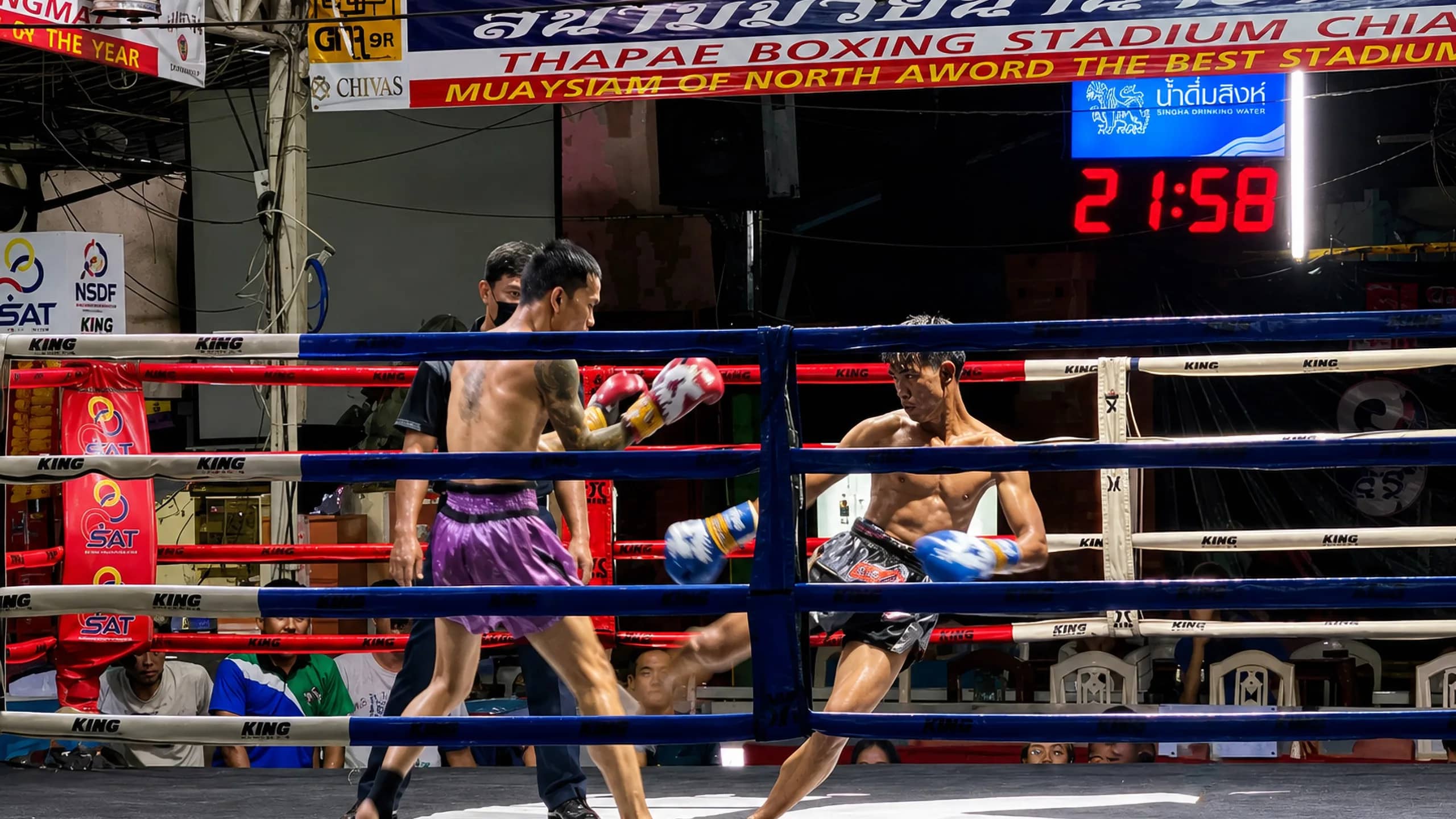 Two Muay Thai fighters in red and blue gloves exchange blows in the ring at Thaphae Stadium