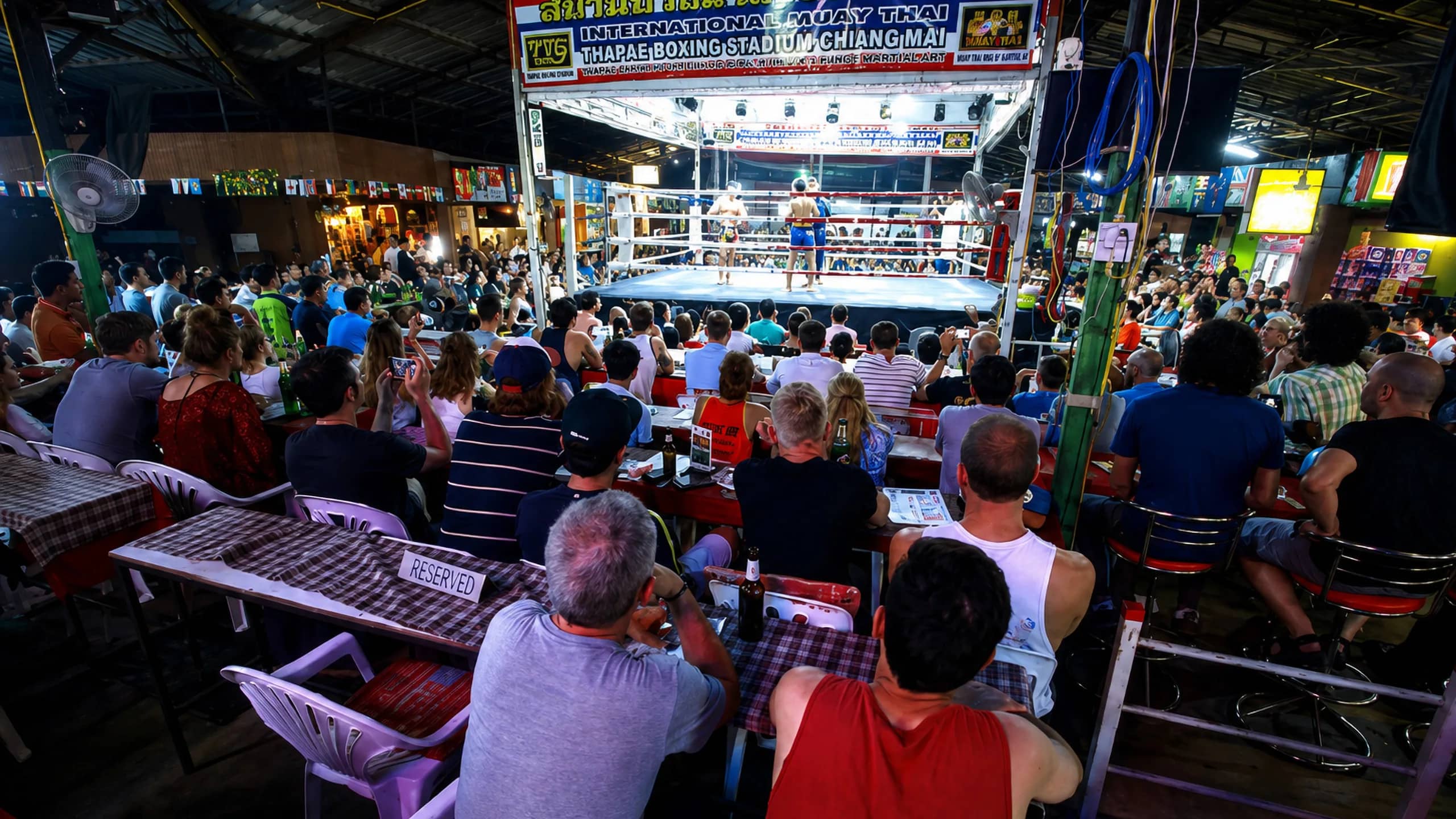 Spectators at tables watch a Muay Thai bout in the brightly lit ring inside Thaphae Stadium