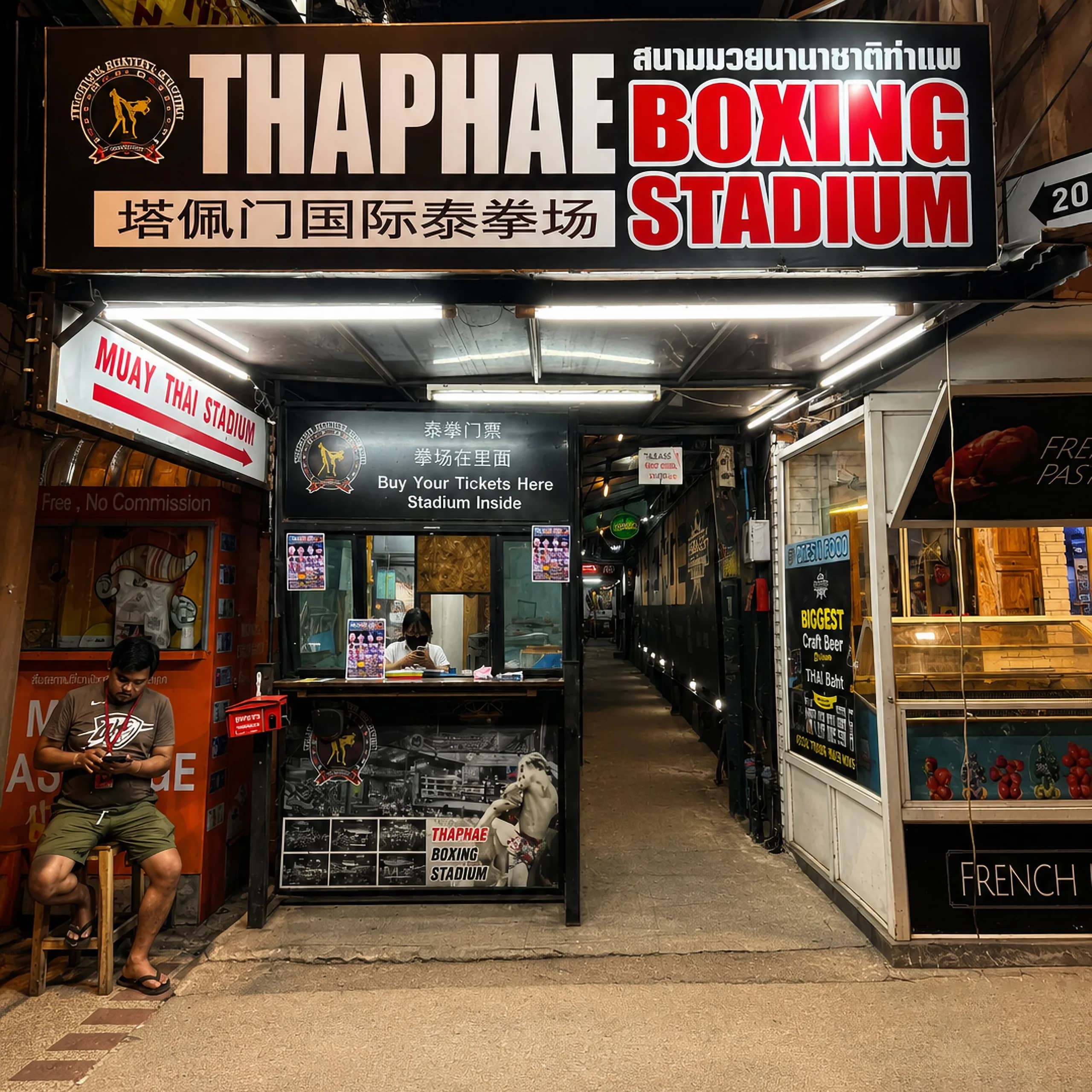 The lit Thaphae Boxing Stadium sign hangs above the ticket booth and the alley entrance at night