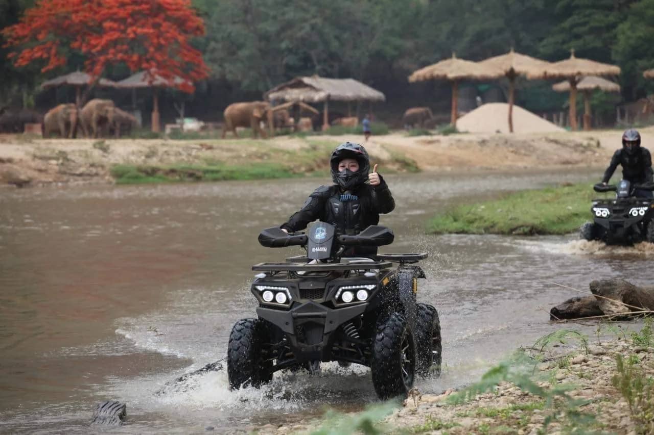 An ATV rider in a helmet and black gear splashes across a shallow river near grazing elephants