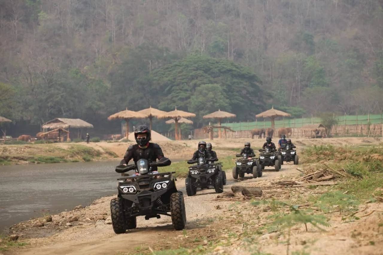 A group of ATV riders follows a sandy riverside track past thatched umbrellas and hills