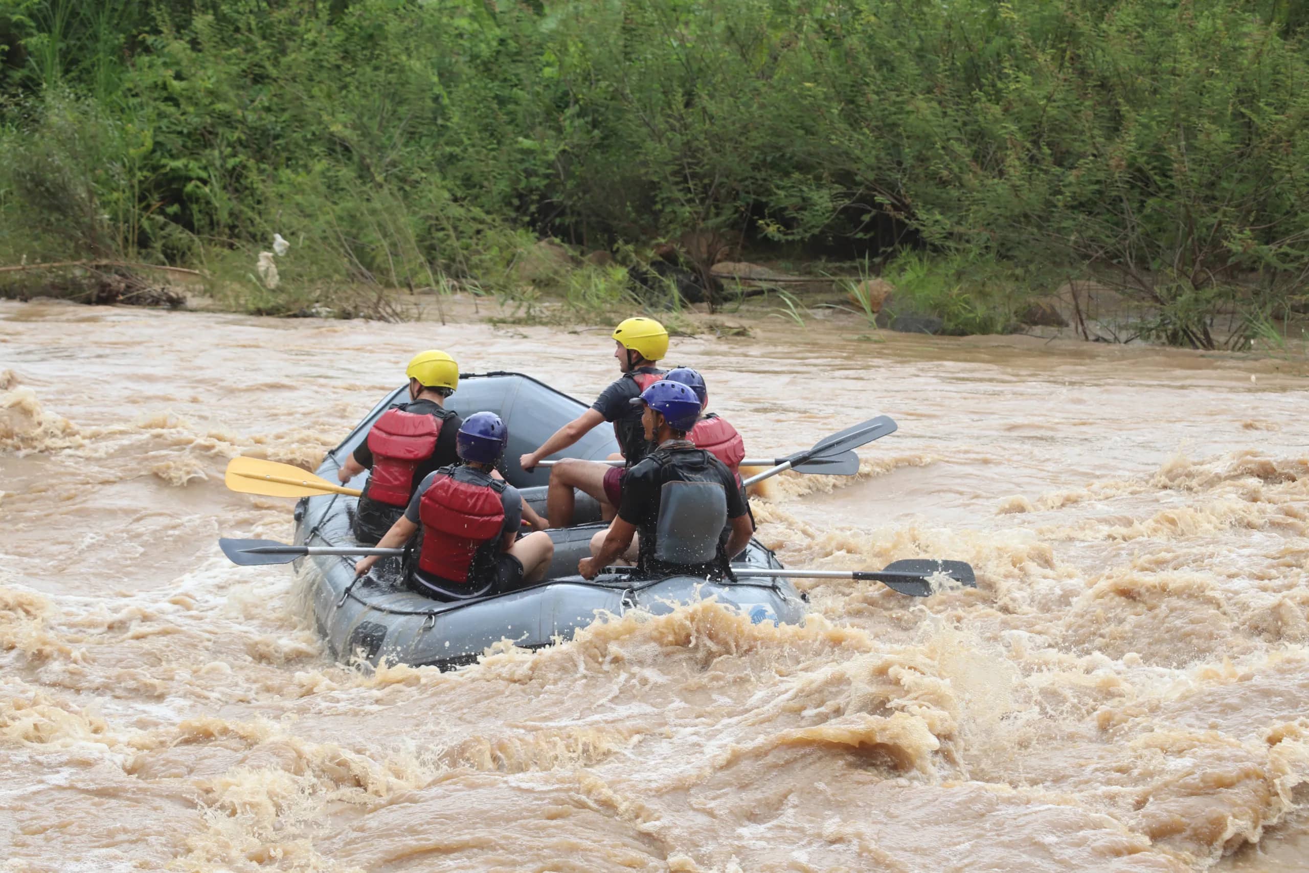 A raft crew in yellow and blue helmets paddles into churning brown rapids beside the jungle