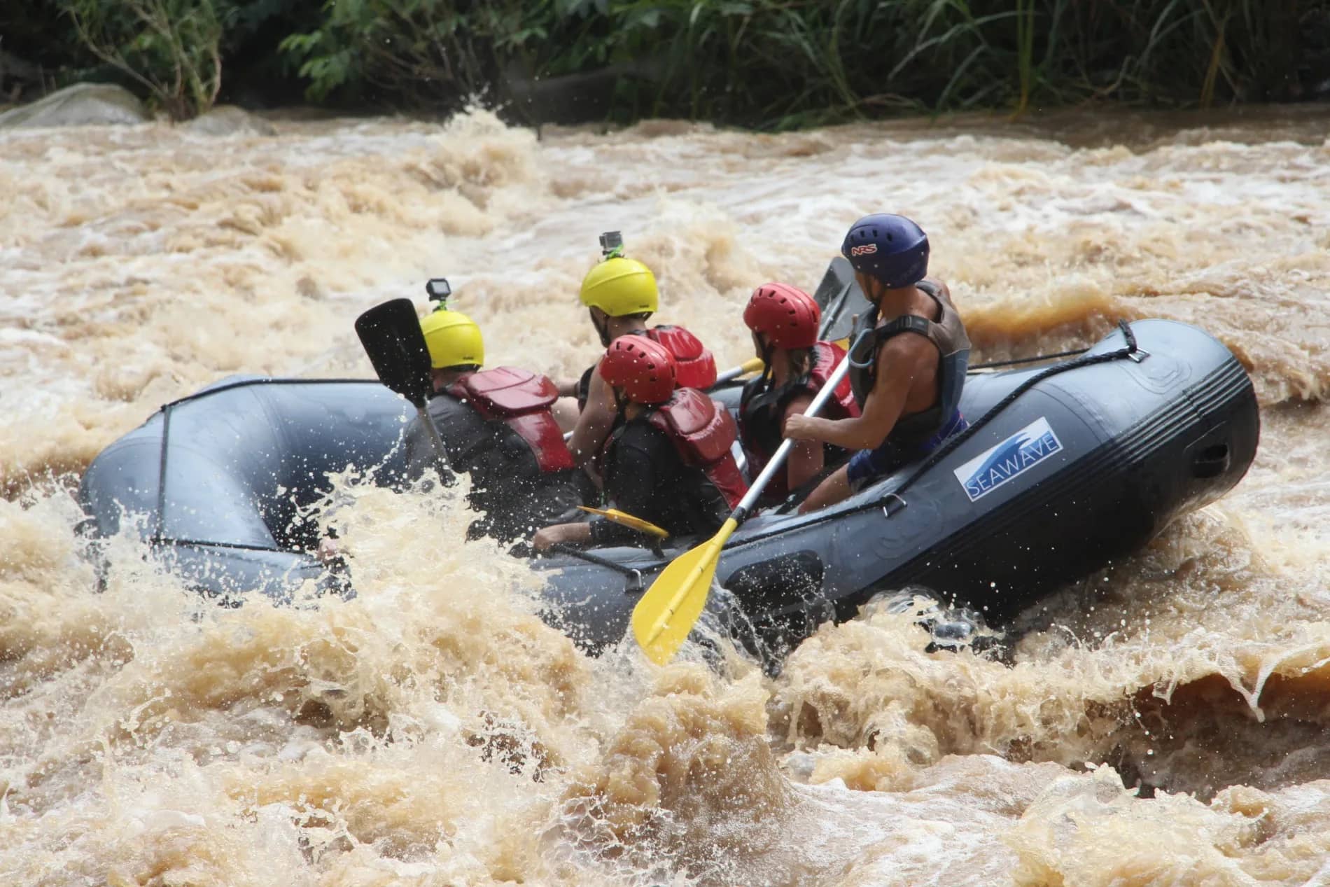 A grey raft with paddlers in helmets and life jackets plunges through foaming brown whitewater