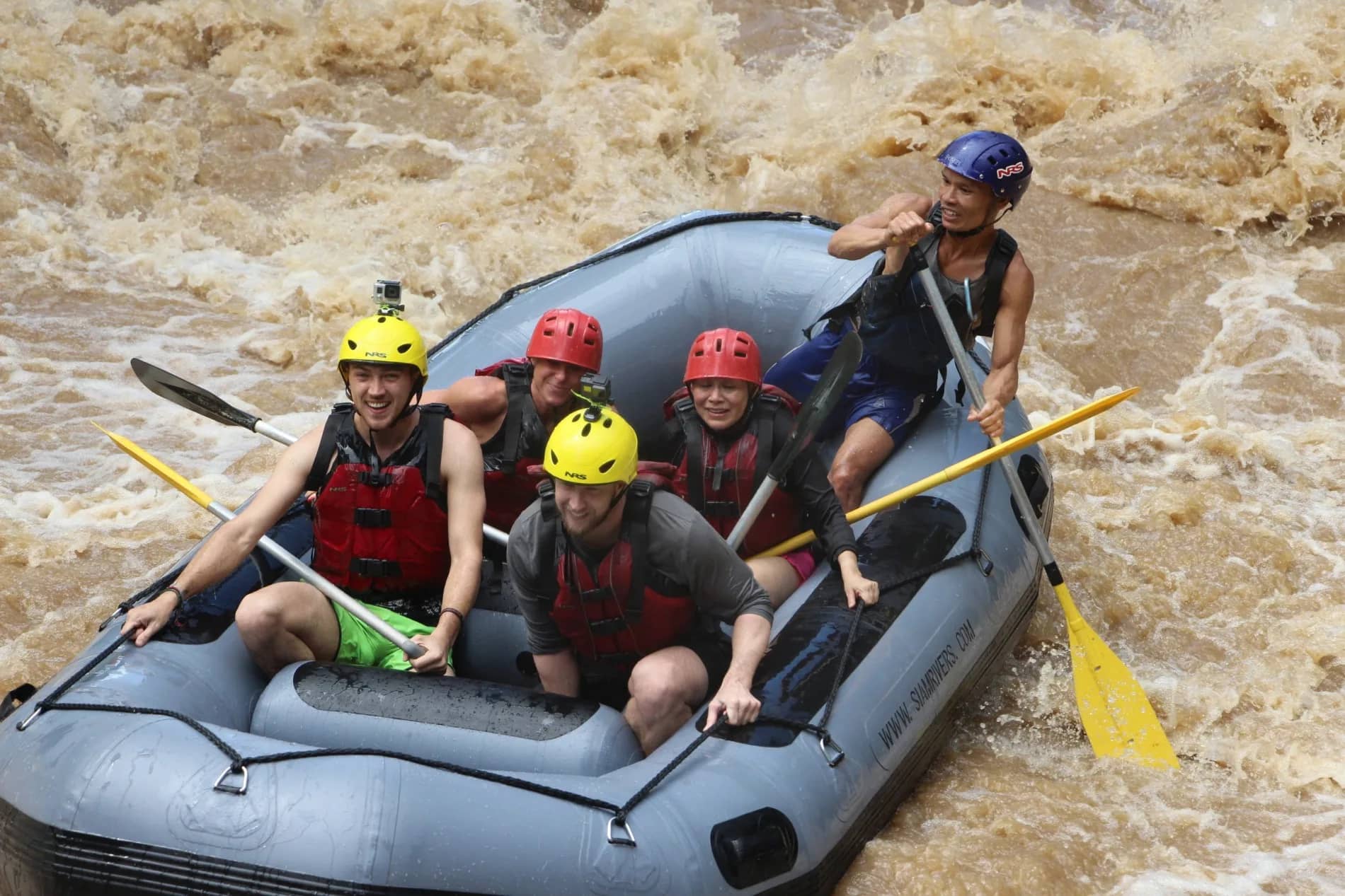Smiling rafters in colourful helmets ride a grey raft through rough brown rapids with their guide