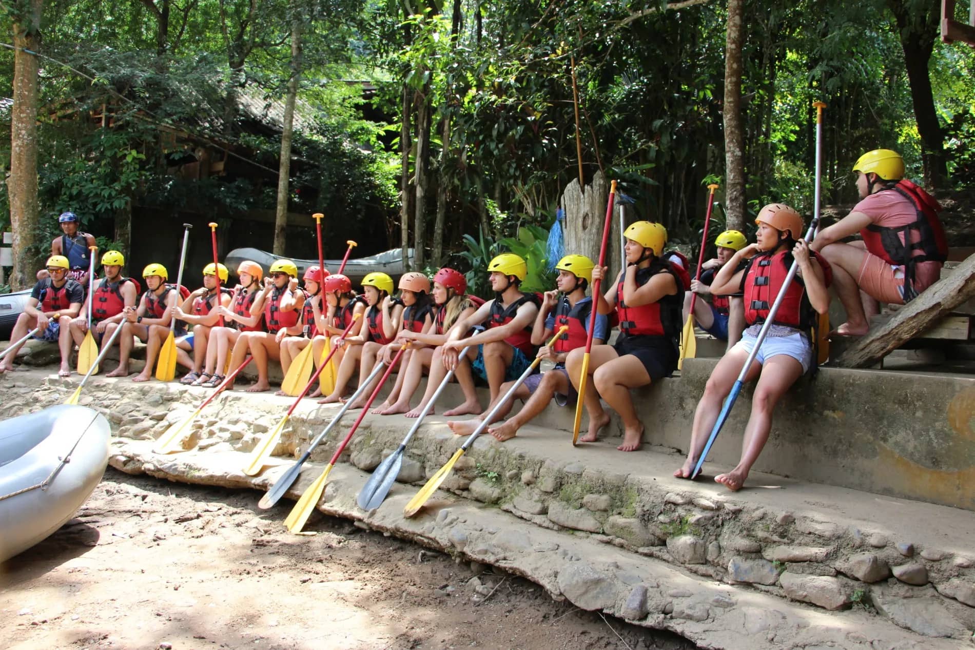 Rafters in helmets and life jackets sit in a long row with paddles on a stone wall