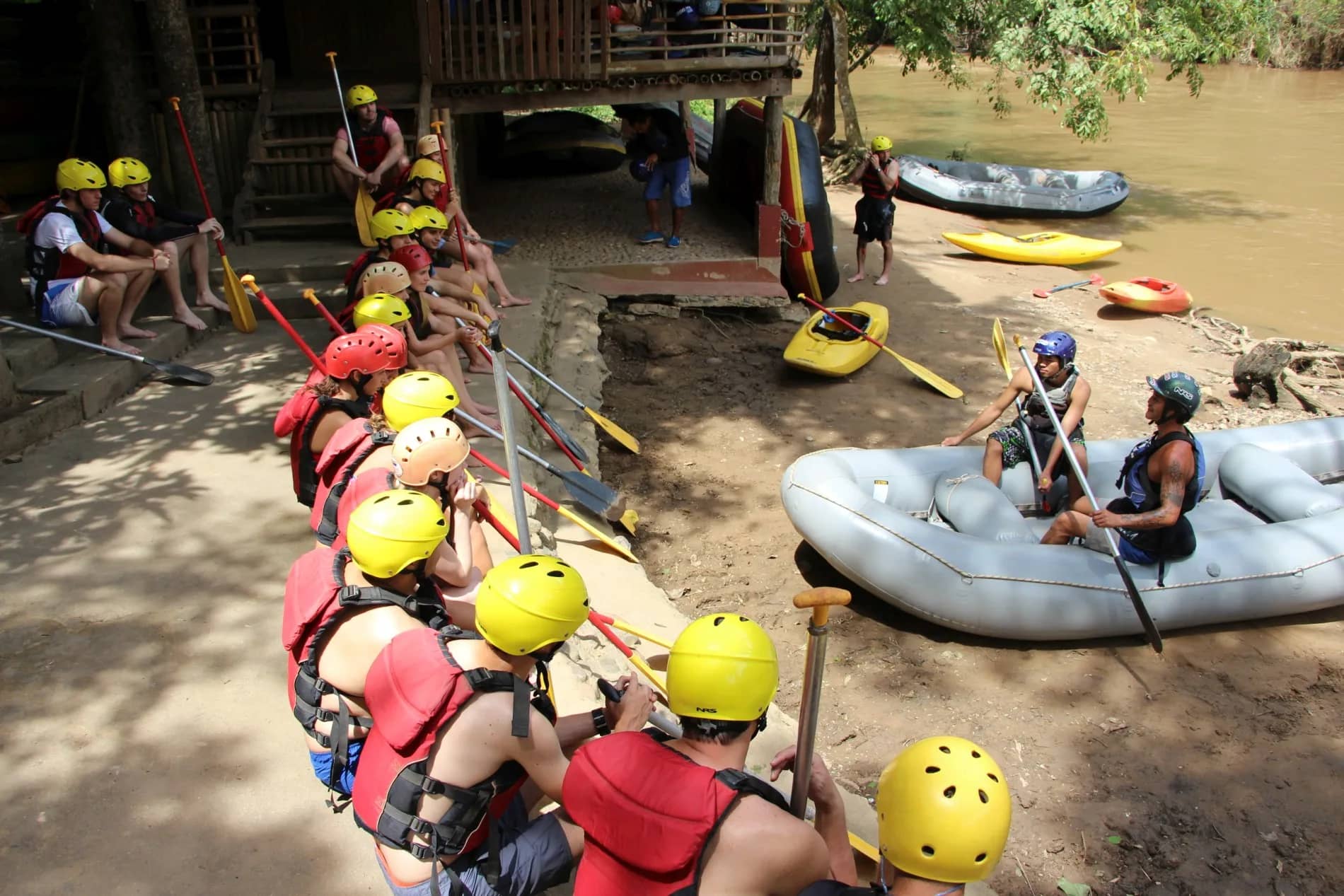 Rafters in yellow helmets sit with paddles beside rafts at the river launch area