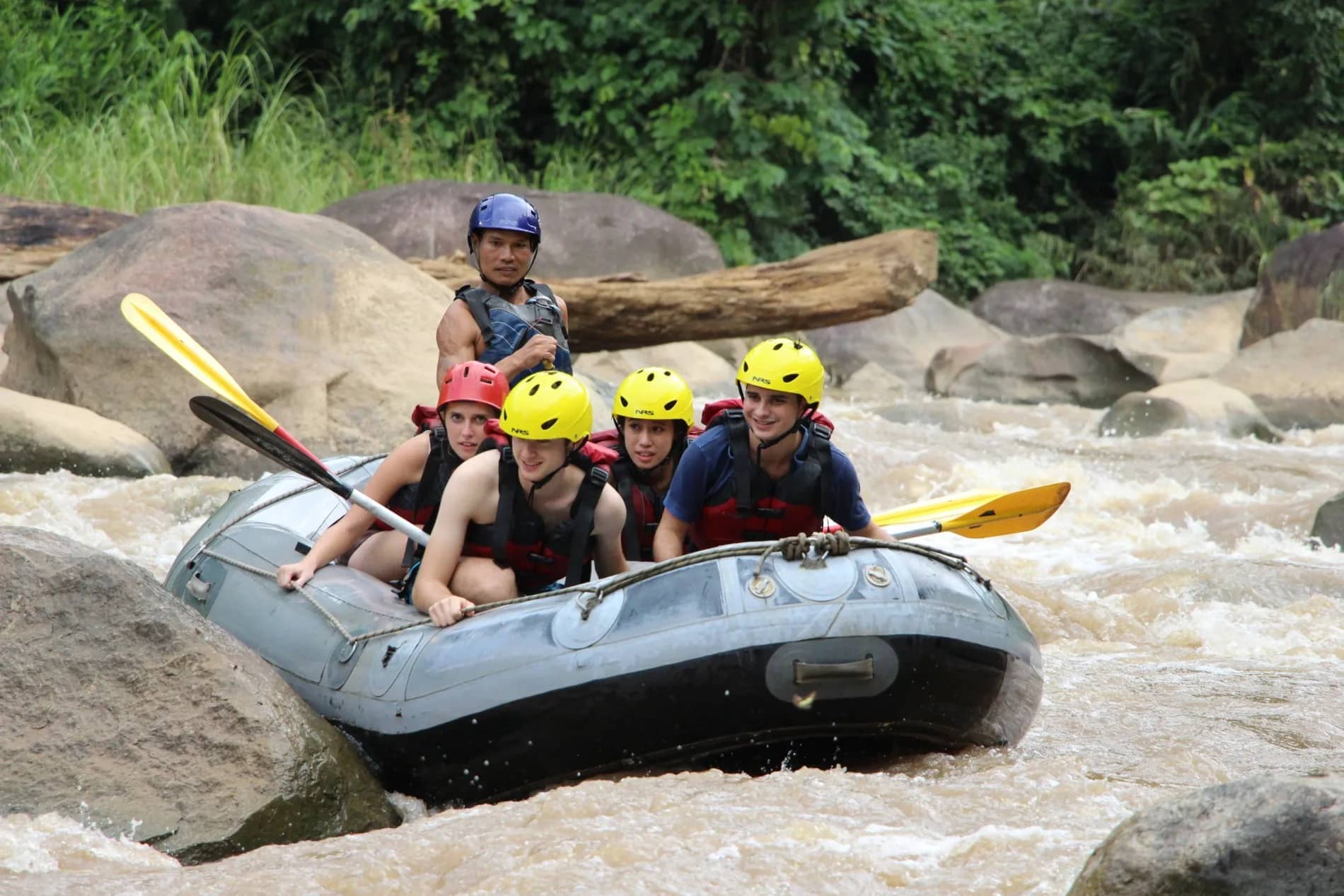 A rafting crew in helmets steers a grey raft past big boulders in fast jungle water