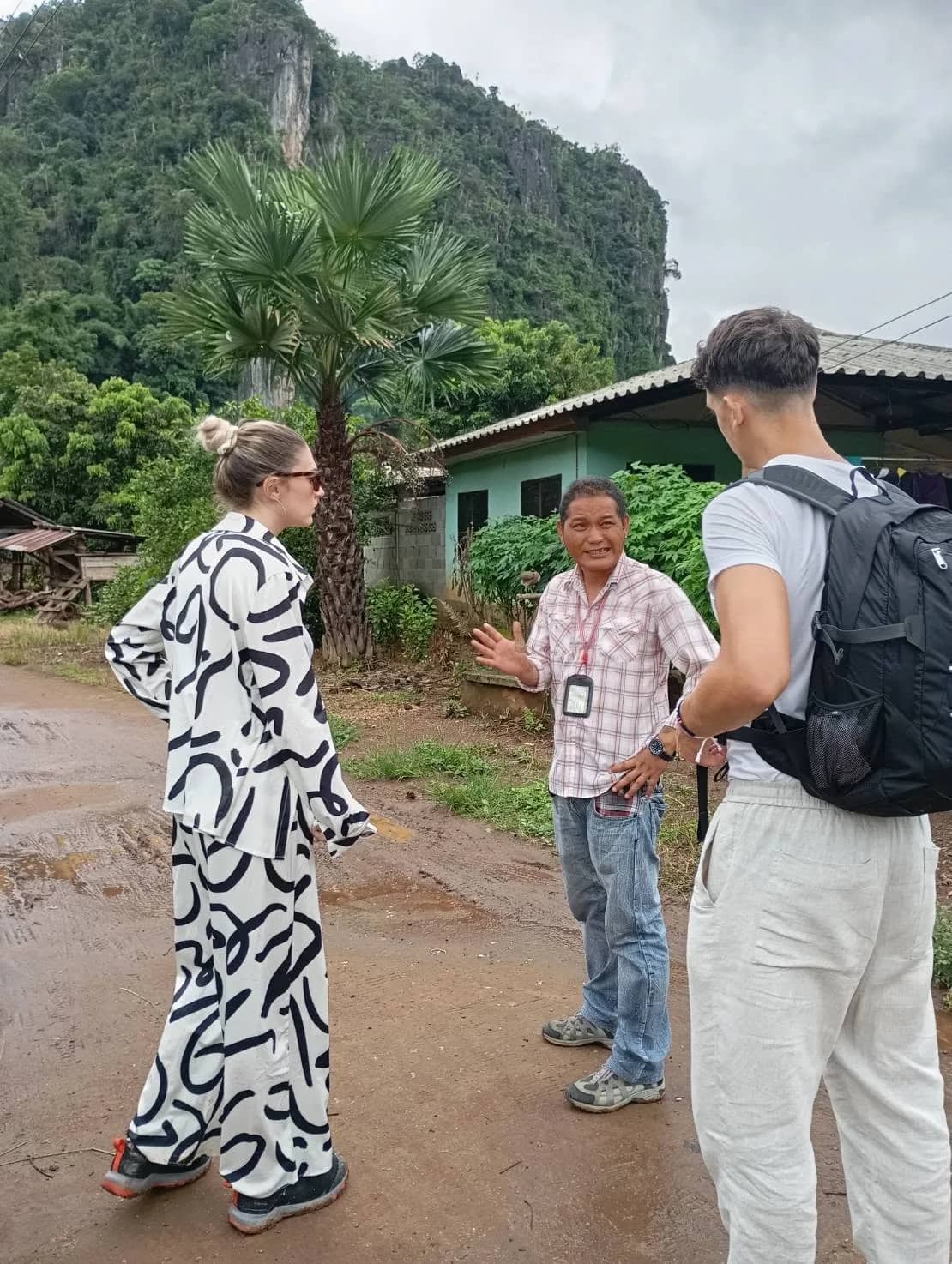Two visitors talk with a local man on a dirt lane in a village below limestone cliffs near Chiang Dao