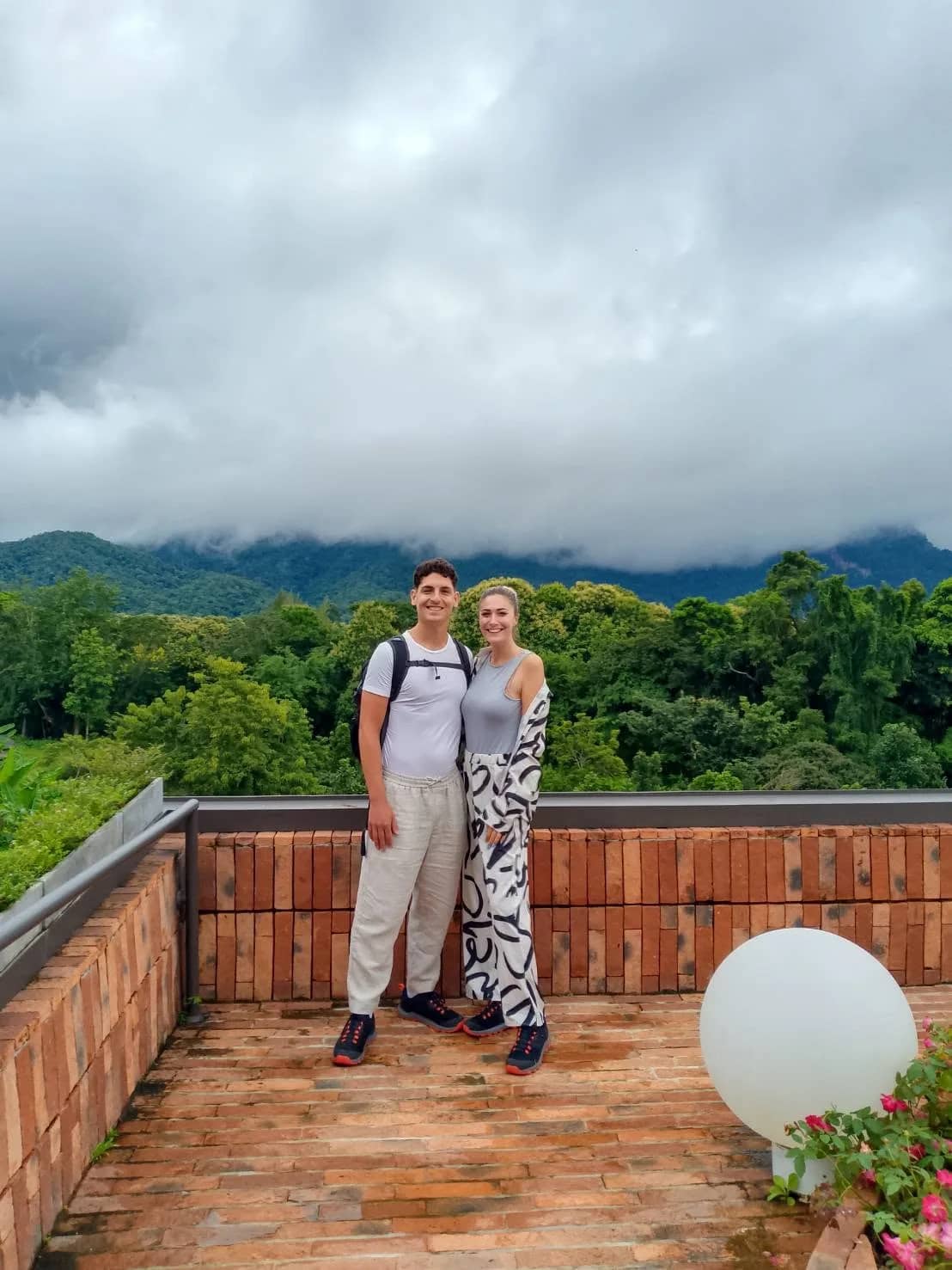 Two visitors stand on a brick rooftop terrace with clouds over the forested Chiang Dao mountains behind
