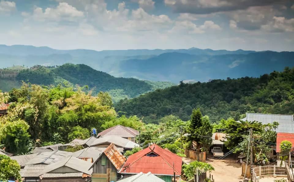 Tin and tiled roofs of a hill tribe village look out over layered green mountains near Chiang Dao