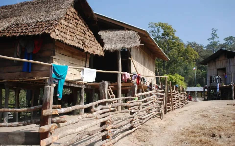 Wooden houses on stilts with thatched roofs line a dirt lane in a hill tribe village