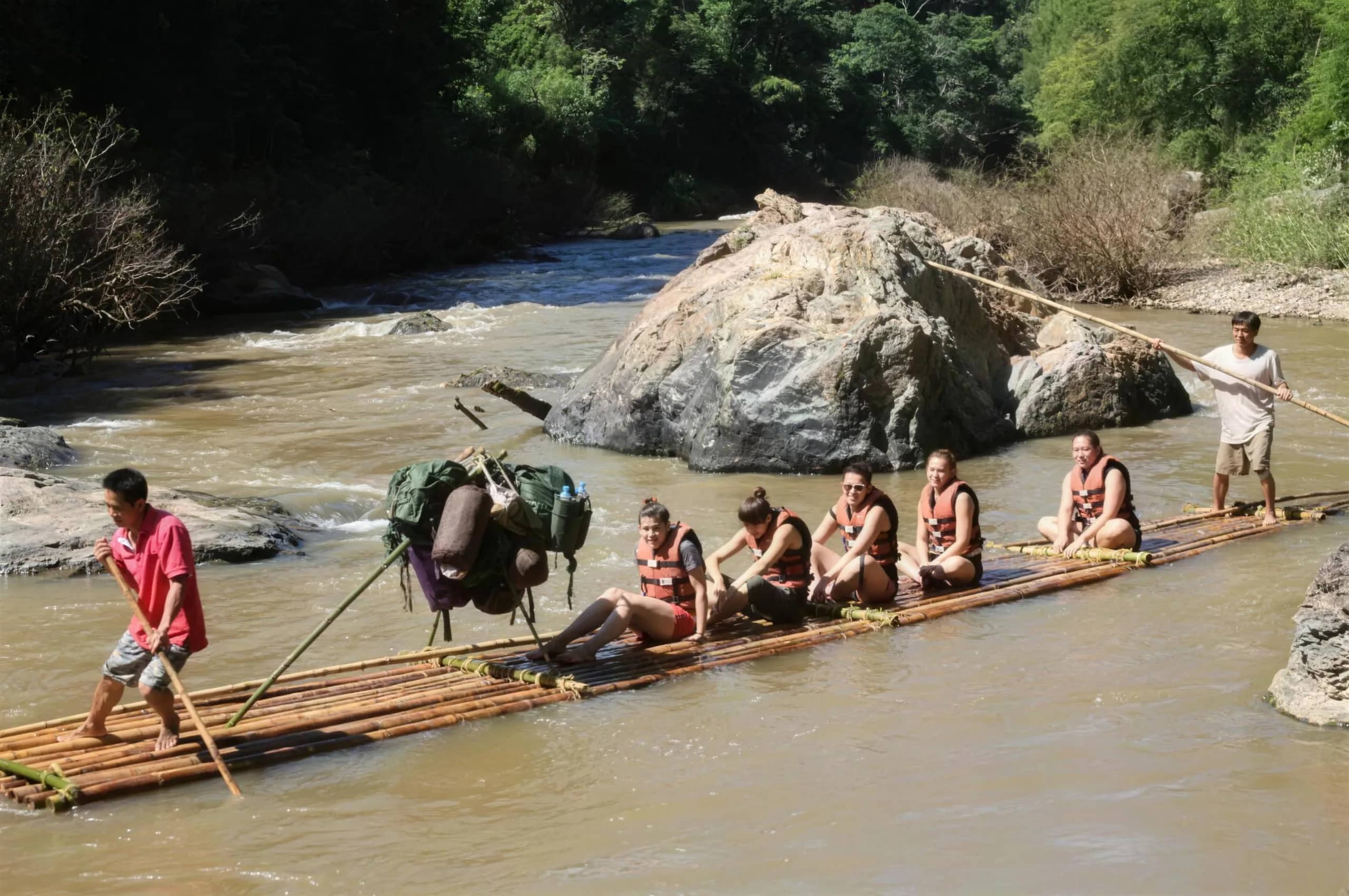 Guests in life jackets sit on a long bamboo raft steered by guides with poles on a brown river