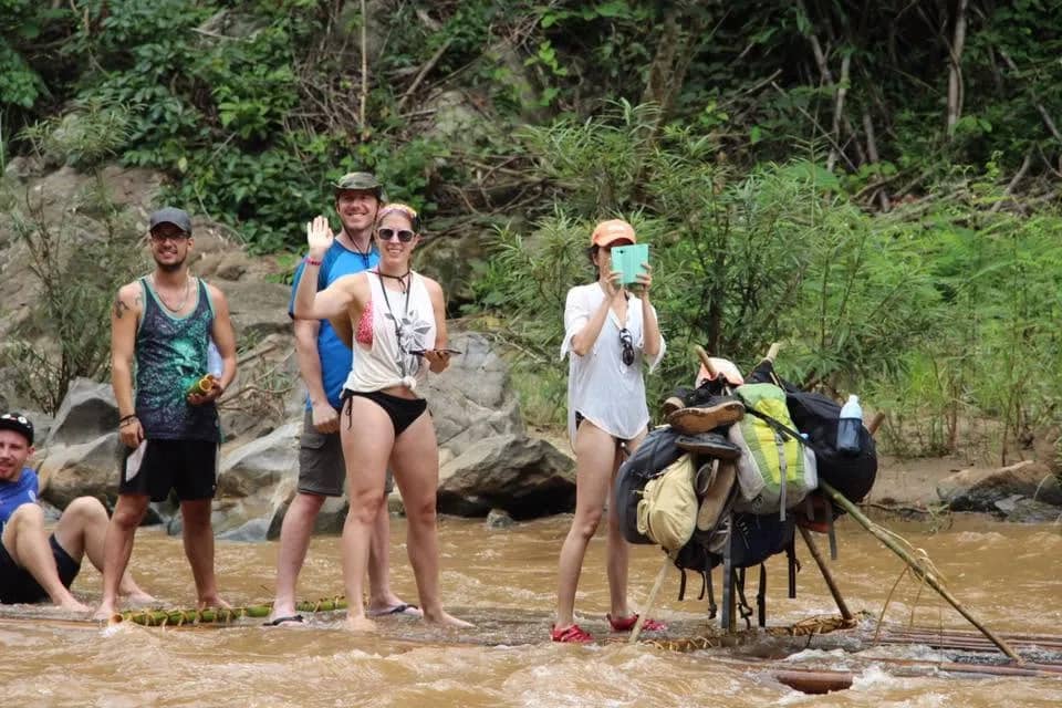 Rafters in swimwear wave from the shallow water beside a bamboo raft loaded with backpacks
