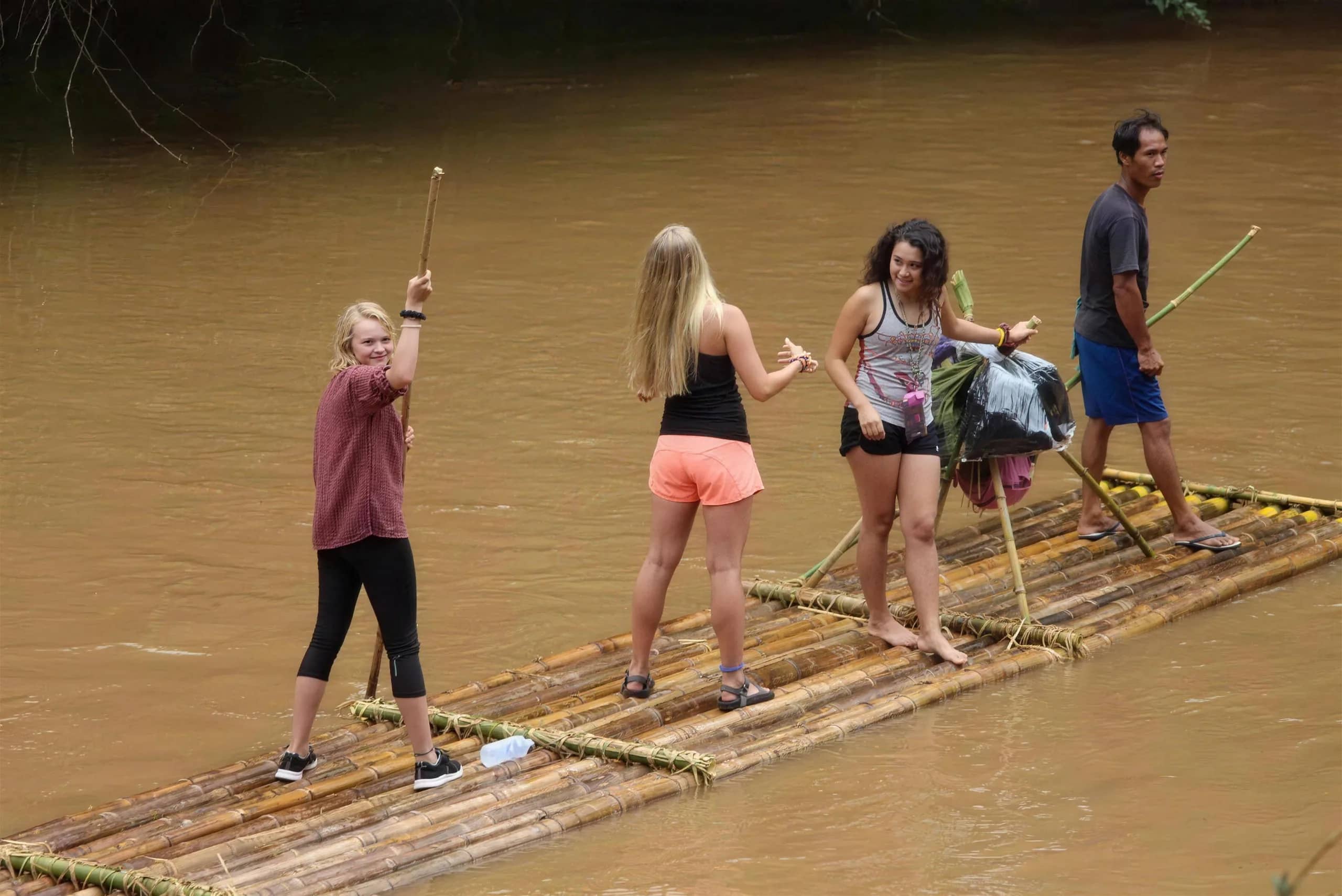 Guests stand on a bamboo raft holding long poles as a guide steers on a muddy river