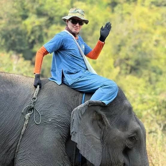 A smiling guest in a blue outfit sits on an elephant's neck and waves
