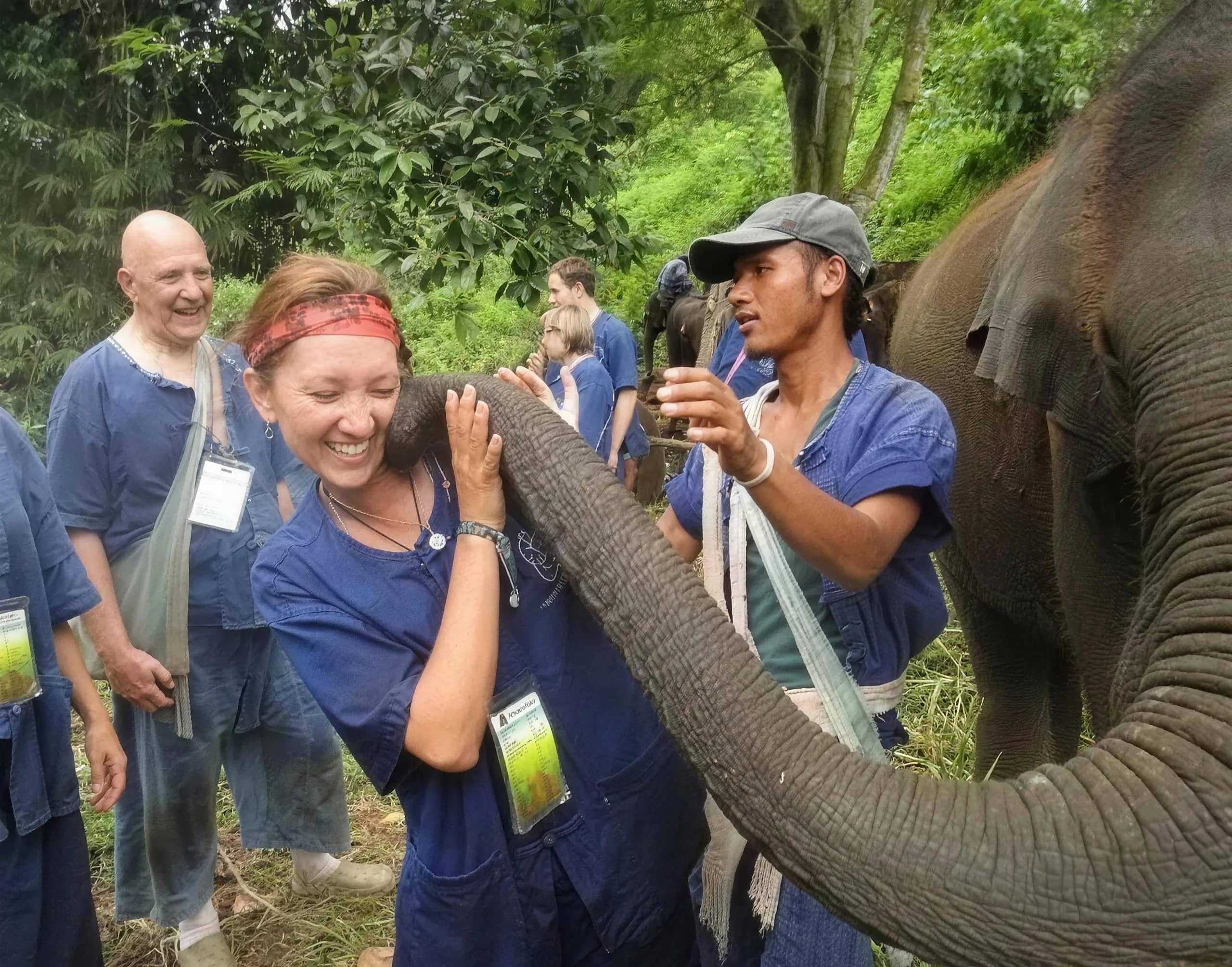 A laughing visitor in a blue shirt is hugged by an elephant's trunk as others watch