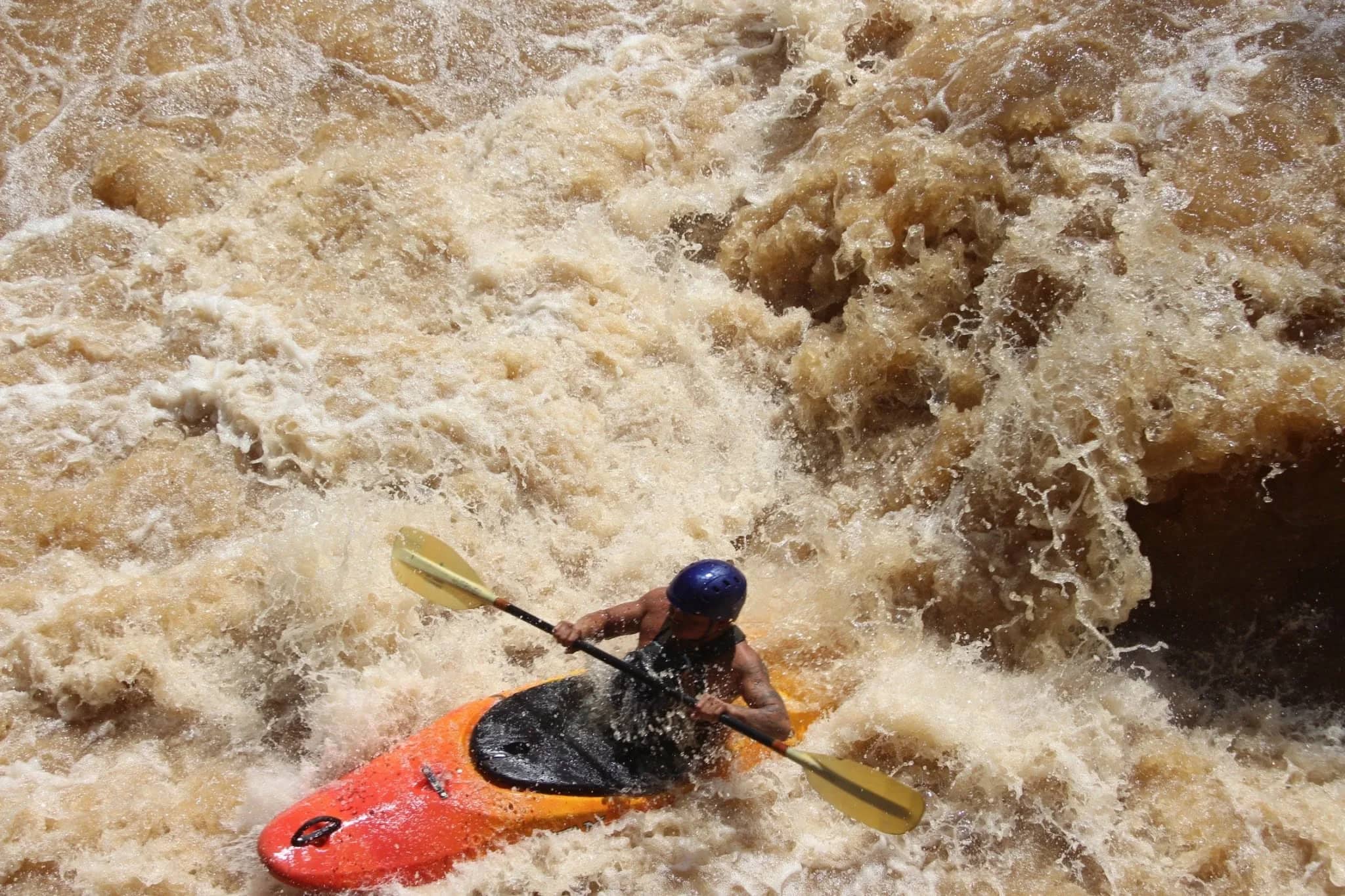 A kayaker in a red boat and helmet paddles through churning brown rapids near Chiang Mai