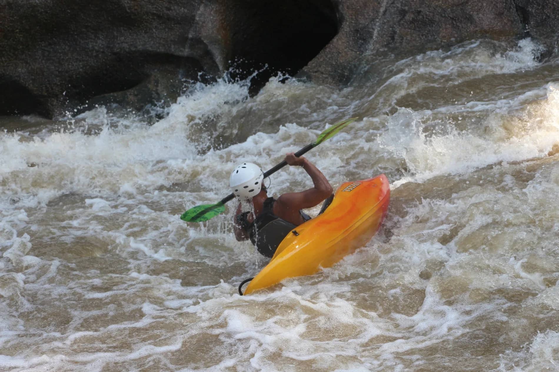 A kayaker in an orange boat braces with the paddle in white rapids below dark rocks