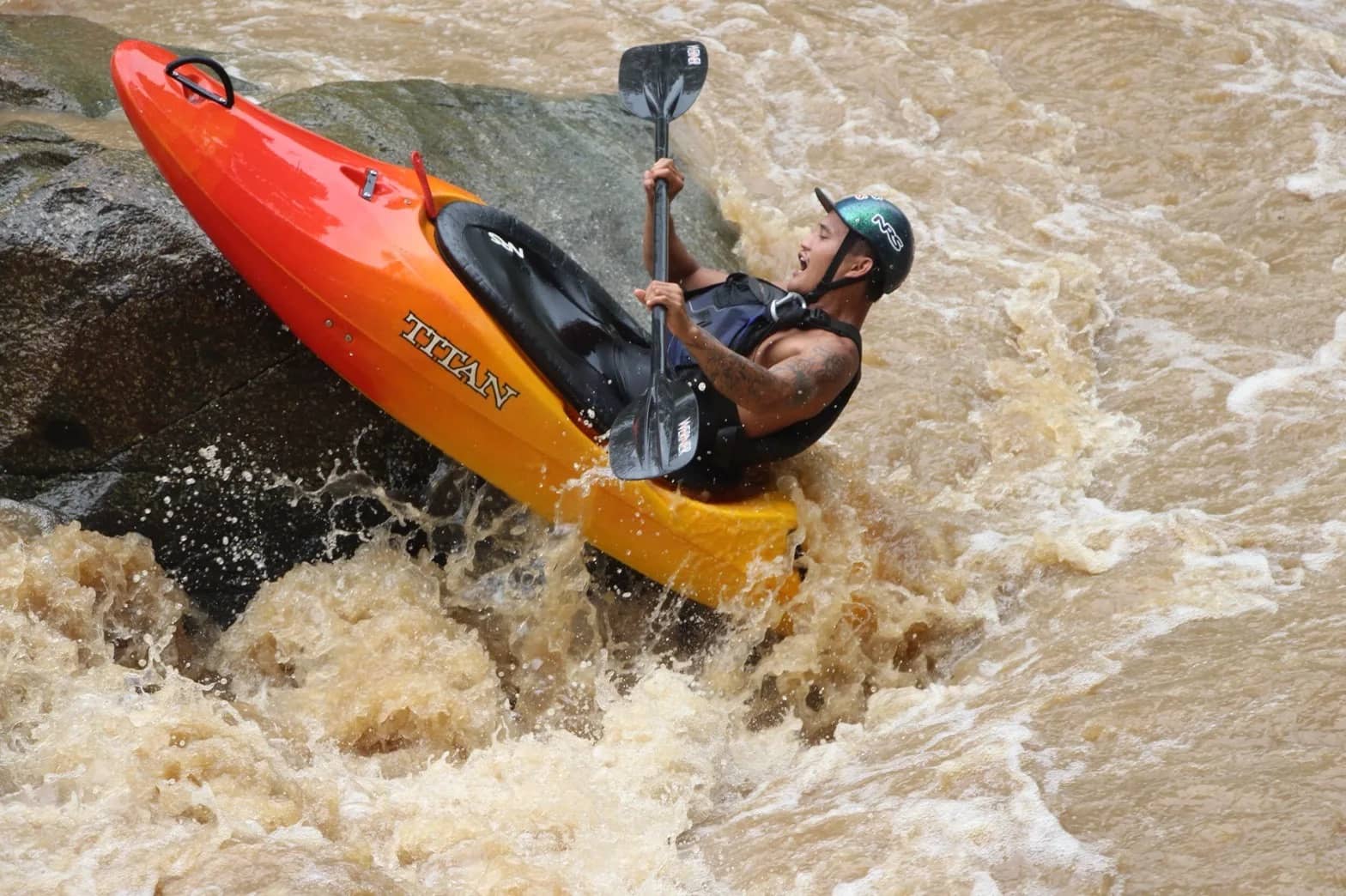 A kayaker steers an orange whitewater kayak steeply off a rock into a fast brown river