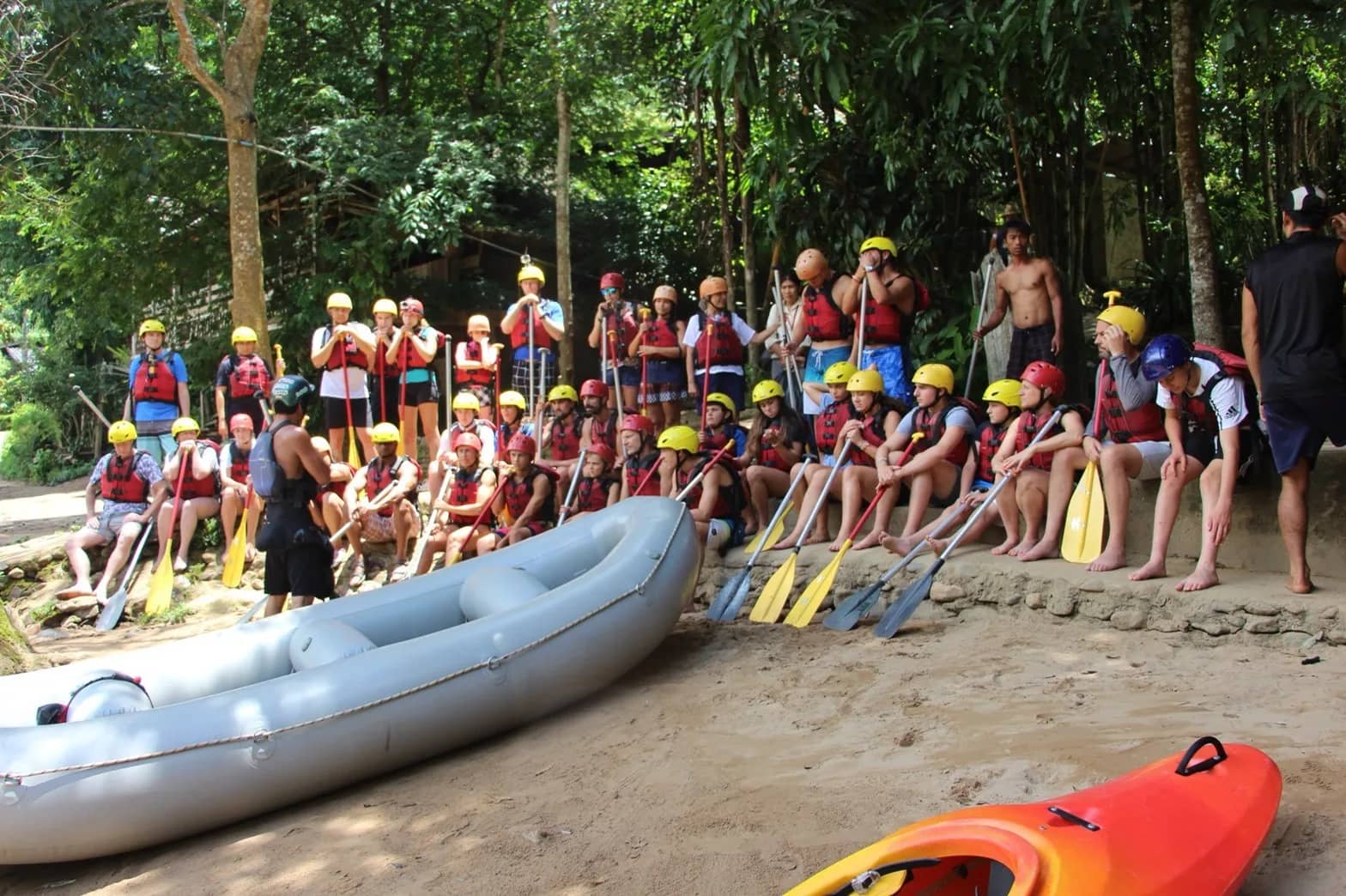 A large group in helmets and life jackets holds paddles beside an inflatable raft before setting off