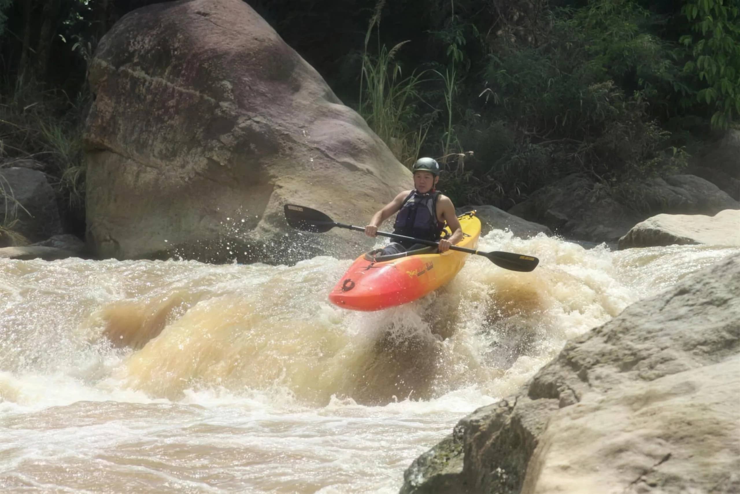 A kayaker in a red boat drops through a foaming rapid between large boulders on a jungle river
