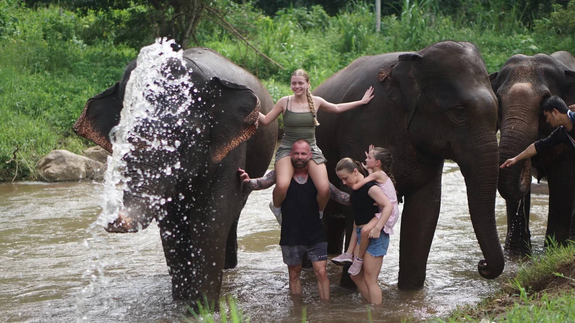 An elephant sprays water from its trunk while visitors stand in the river among the herd.
