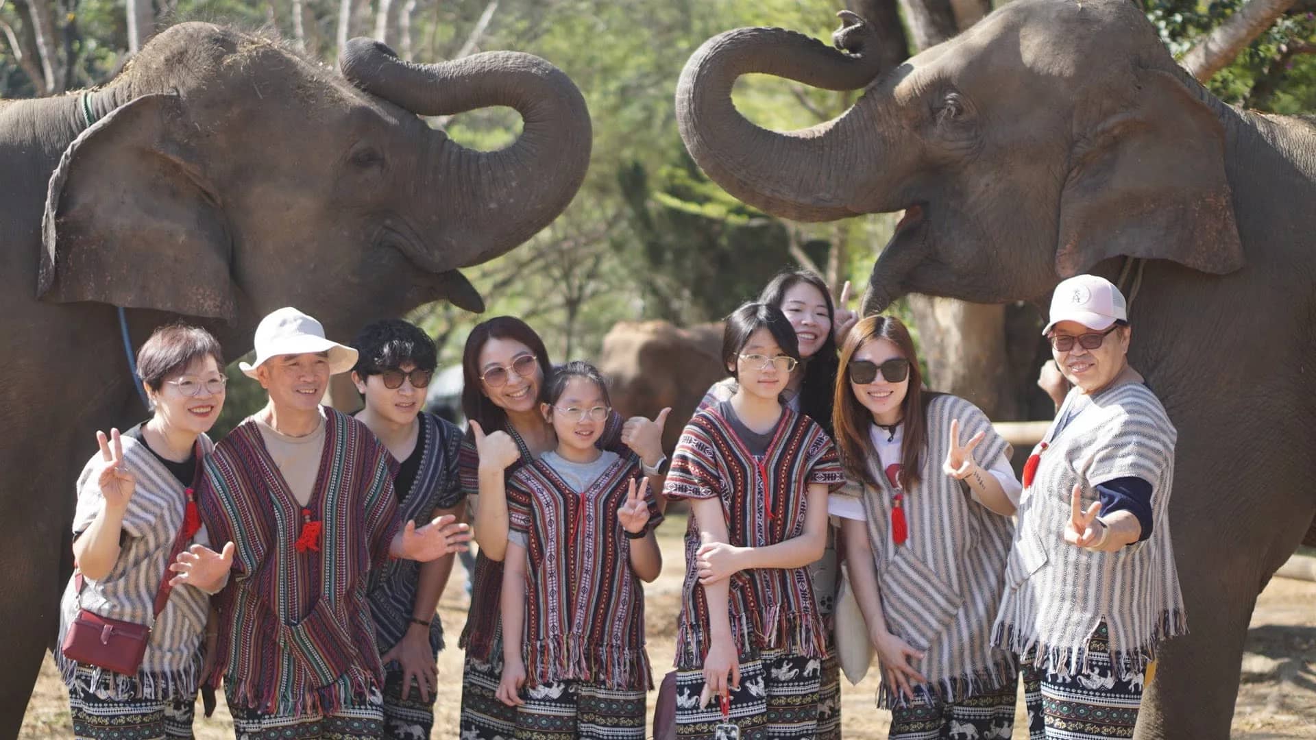 A group in striped woven tops poses between two elephants curling their trunks overhead.