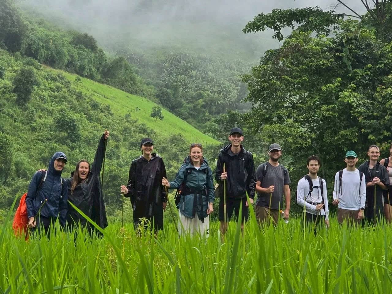 A smiling group of trekkers stands in a green rice field below misty hills