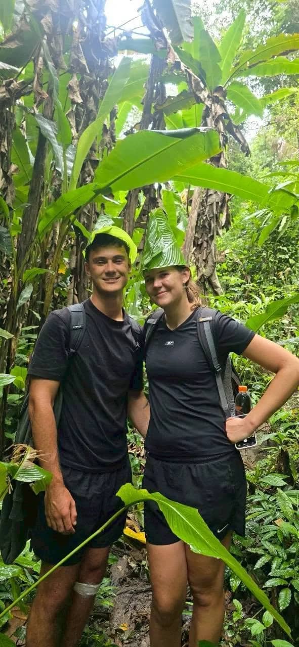 Two hikers with backpacks pose on a jungle trail surrounded by large banana leaves